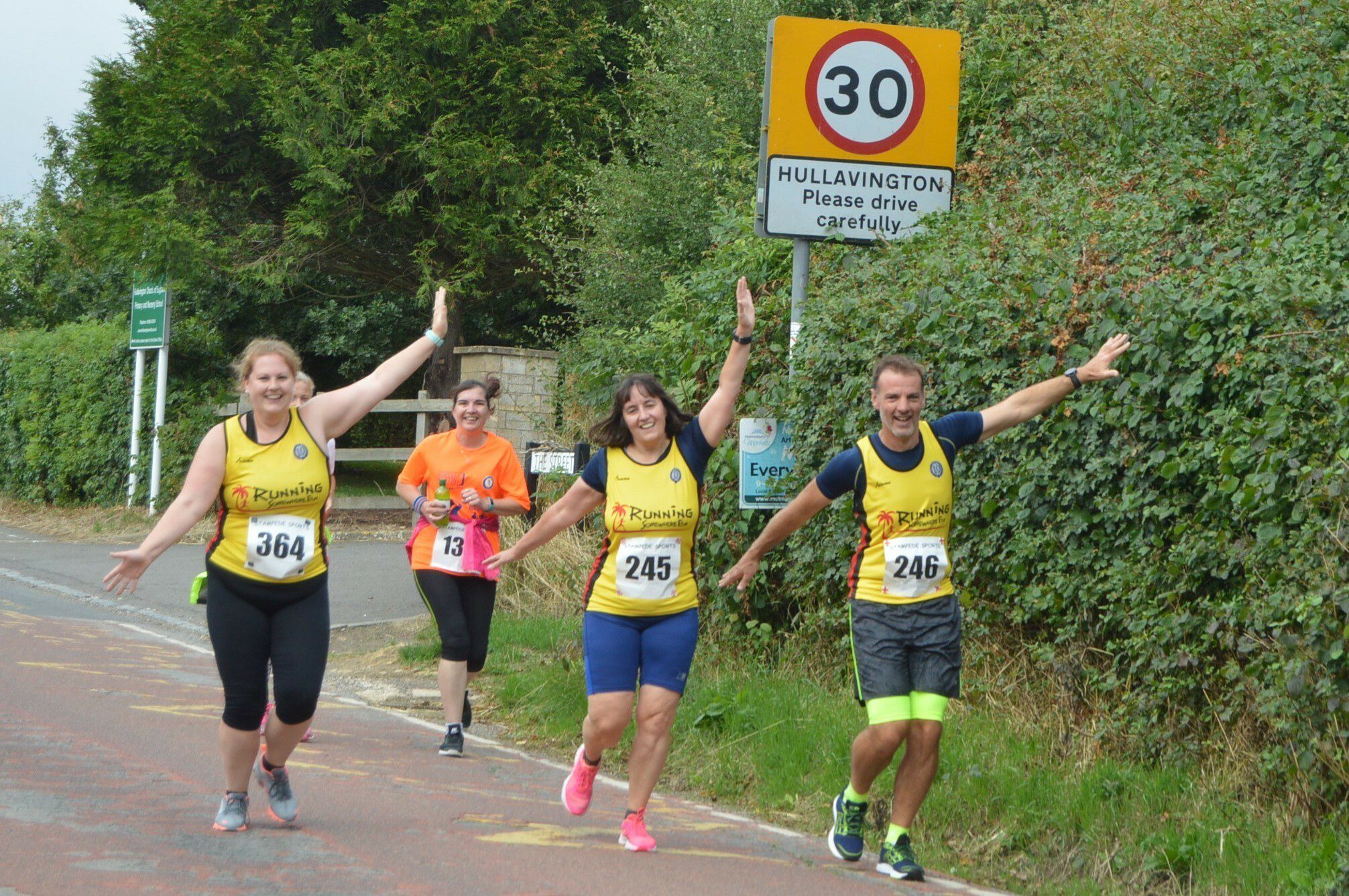 Four runners participate in a race, with three in yellow jerseys (two women, one man) and one in an orange jersey (woman). All are smiling, raising their arms in celebration. A 30 mph speed limit sign and a "Hullavington, Please drive carefully" sign are visible in the background.