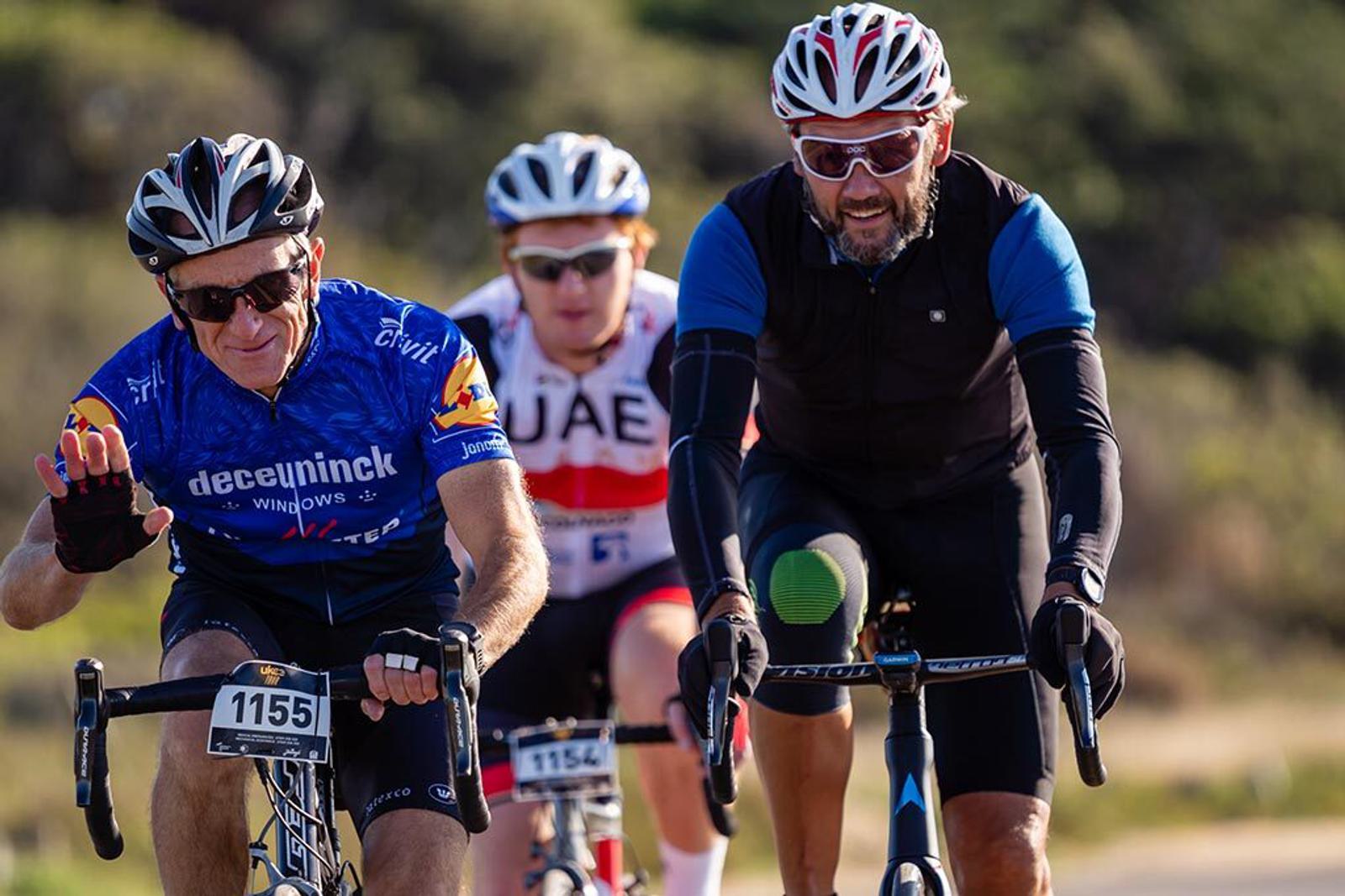 Three cyclists are riding on a road during the day. The cyclist on the left is wearing a blue jersey and waving, the one in the middle is wearing a white and red jersey, and the cyclist on the right is in a black outfit with green knee padding and sporting sunglasses and a beard.