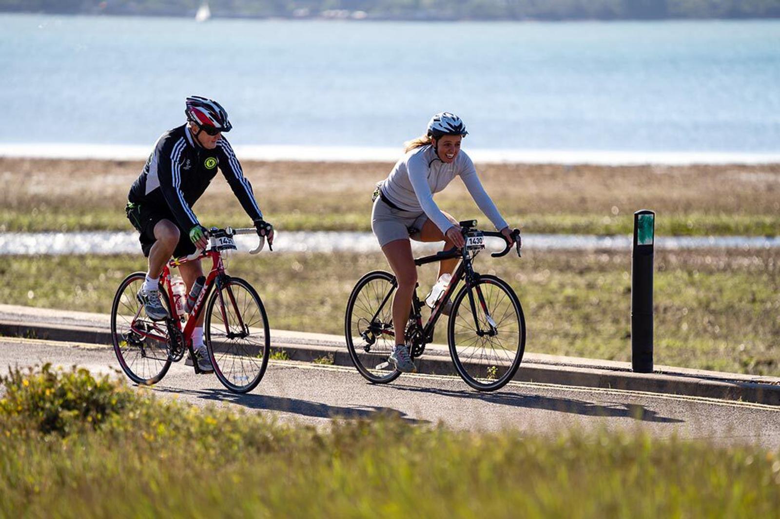 Two cyclists ride along a coastal path on a sunny day. The person on the left wears a black long-sleeve jersey and a helmet, while the person on the right wears a white long-sleeve shirt and a helmet. The background features a serene body of water and greenery.