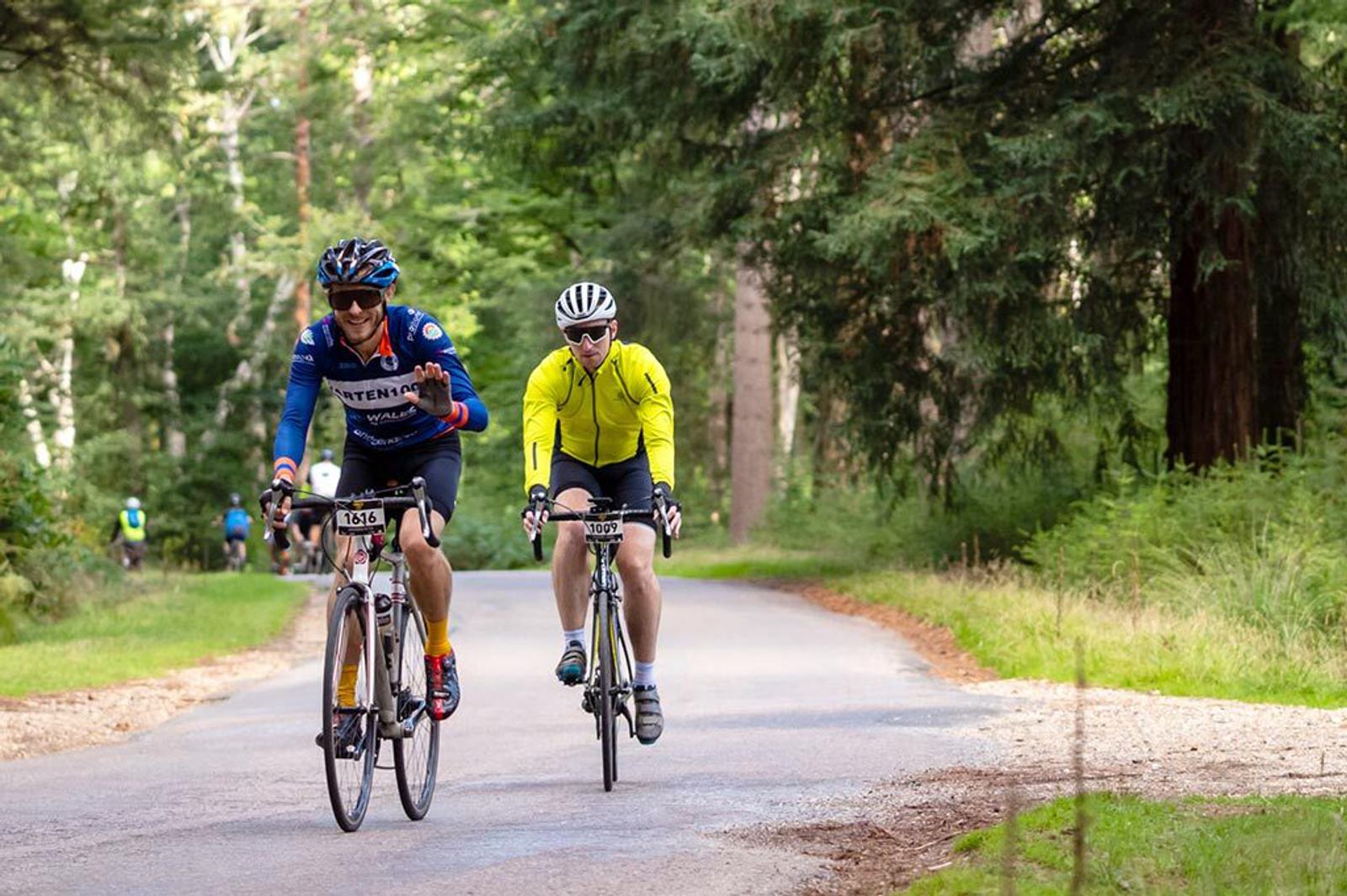 Two cyclists ride on a forested road. The cyclist on the left, wearing a blue and red jersey and a blue helmet, holds up a hand signal. The cyclist on the right, in a yellow jacket and white helmet, pedals alongside. Green trees and foliage surround the path.