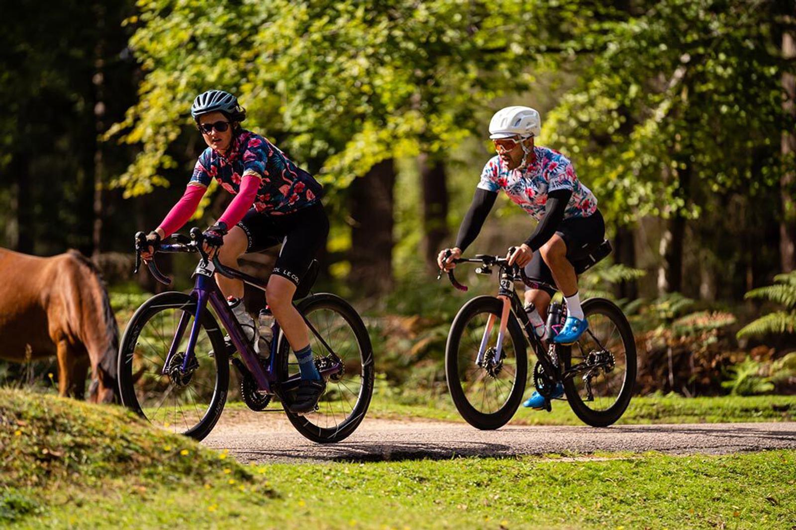 Two cyclists, wearing colorful jerseys and helmets, ride on a paved path through a lush green park with trees in the background. The cyclist on the left rides ahead, while a horse grazes nearby on the left side of the image.