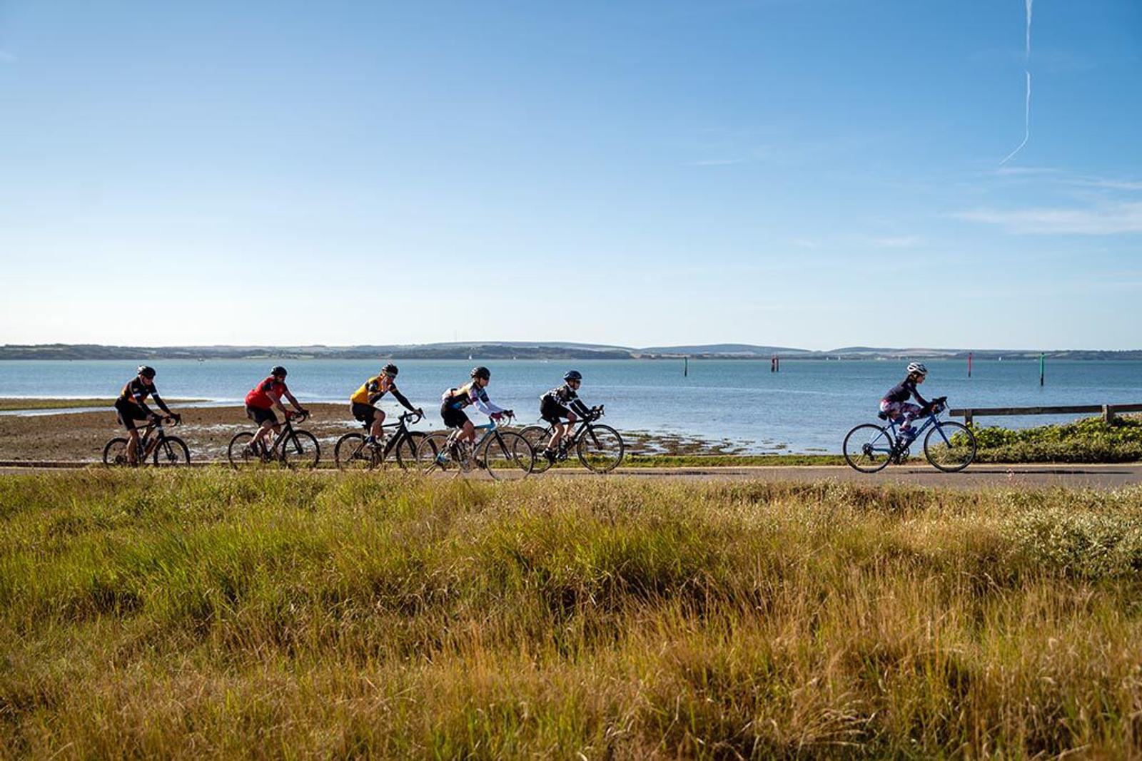 A group of six cyclists rides along a paved path near a body of water under a clear blue sky. The foreground features tall grass, and the background includes water with small poles and a distant shoreline. The cyclists are dressed in colorful athletic gear.