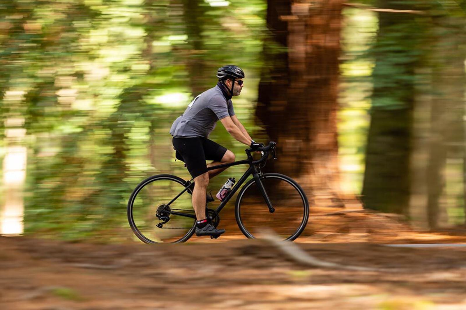 A cyclist wearing a helmet, sunglasses, and cycling gear rides a black bike through a forest with tall trees. The background is motion blurred, giving a sense of speed and movement. The rider has a water bottle attached to the bike frame.