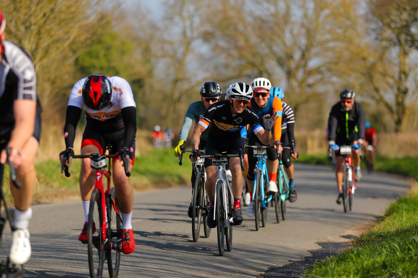 A group of cyclists ride on a rural road with trees in the background. The lead cyclist is focused ahead while wearing a black and white jersey, and others follow closely behind. The early spring foliage and clear skies provide a picturesque setting.