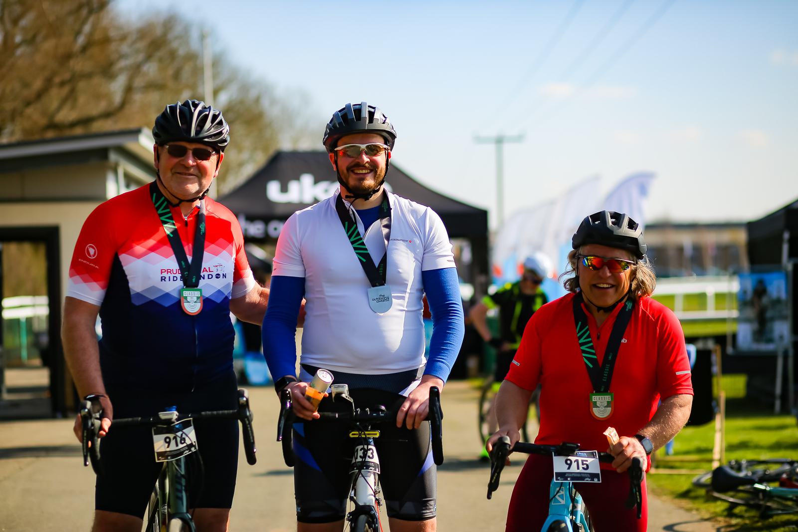 Three individuals in cycling gear are posing with their bikes and wearing medals around their necks. They are outdoors, on a sunny day, with tents and other cyclists visible in the background. The cyclists are smiling and seem to have participated in a biking event.