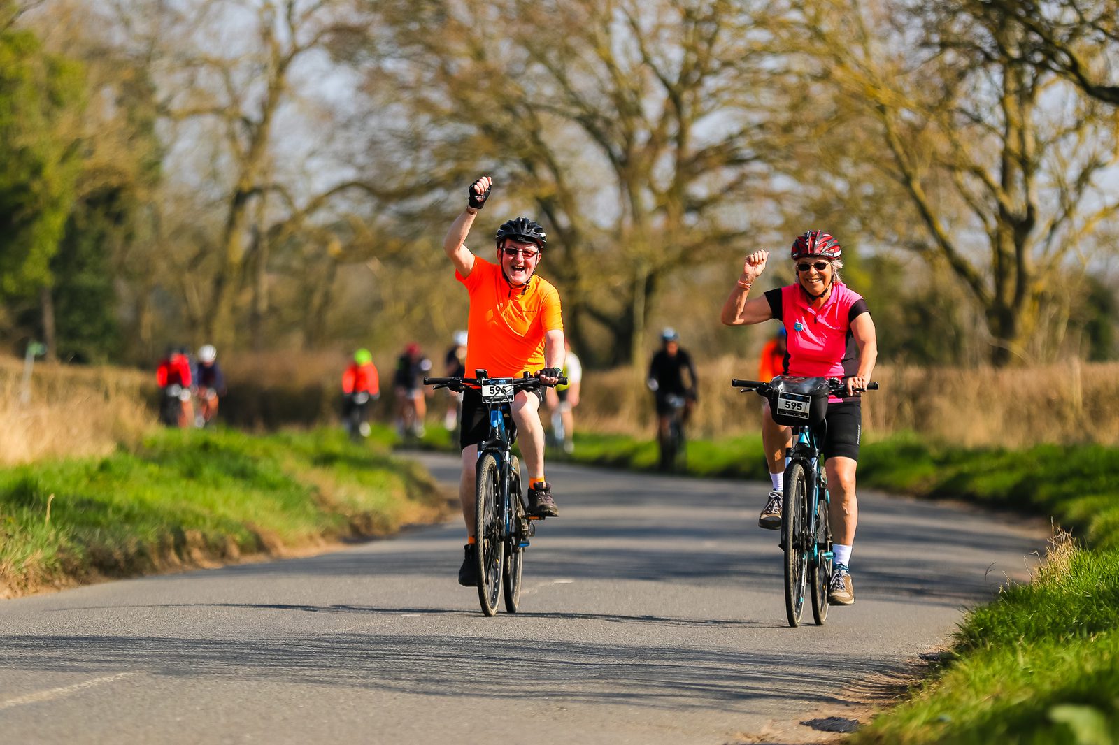 Two cyclists raise their arms in celebration while riding along a scenic, tree-lined rural road. They wear brightly colored gear — one in orange and the other in pink. Both are smiling, with other cyclists visible in the background. The weather is clear and sunny.