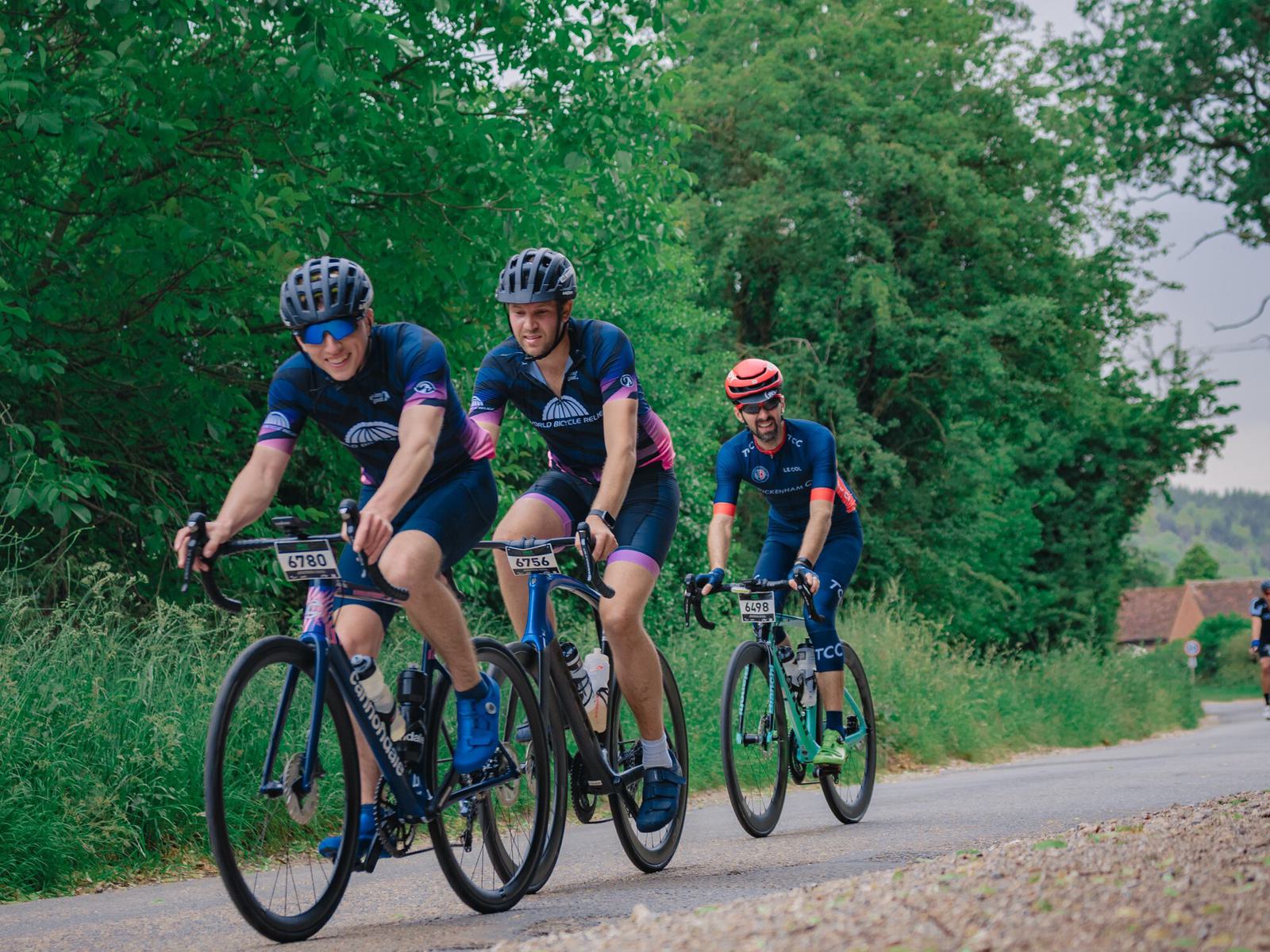 Three cyclists, wearing blue and pink cycling gear and helmets, ride on a paved road lined with greenery. Two cyclists in the lead are side by side, while one follows closely behind, all looking focused and determined. Trees and bushes create a lush backdrop.