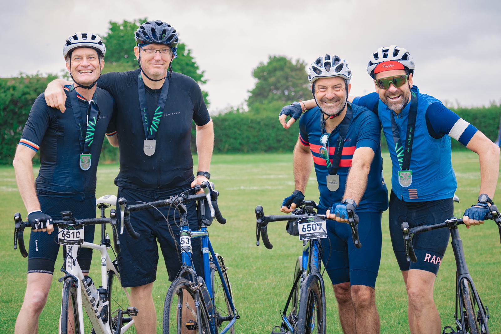 Four men wearing cycling gear and helmets pose with their bicycles on a grassy field. They are smiling and have their arms around each other. The weather is overcast, and they each have numbered race bibs on their bikes.