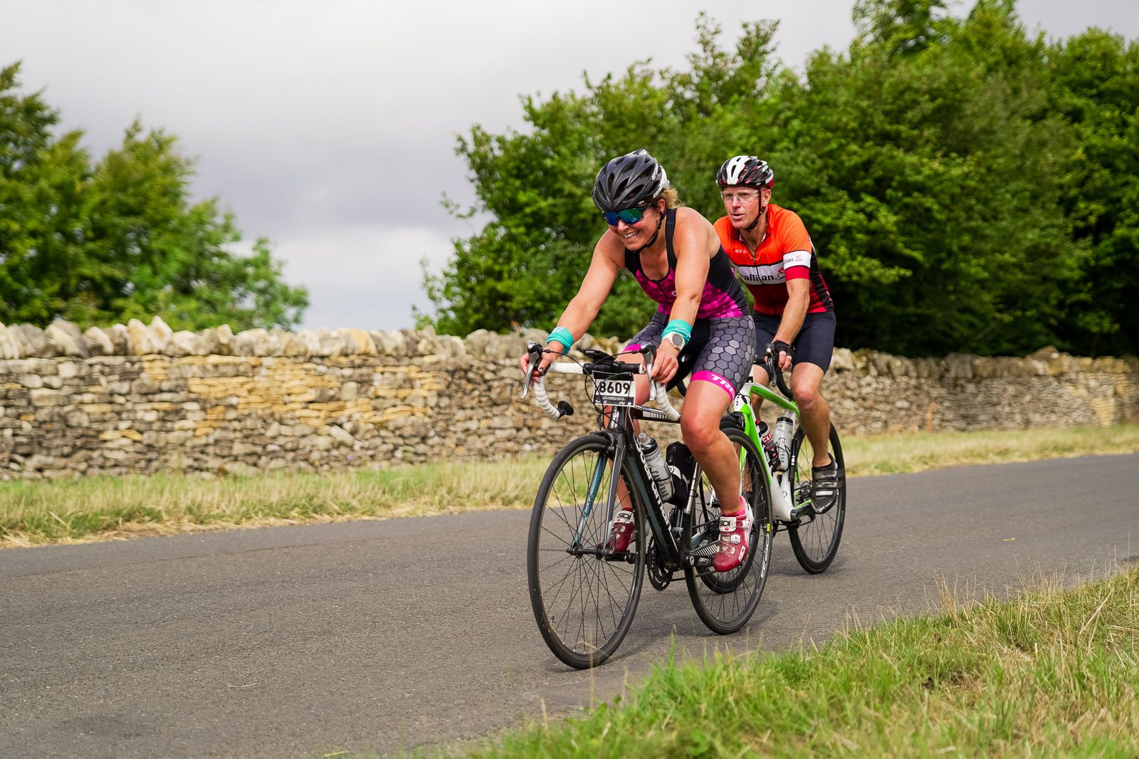 Two cyclists are riding bikes on a paved road with lush greenery and a stone wall in the background. The leading cyclist wears a black helmet, dark sunglasses, and a pink and purple outfit. The cyclist behind them wears a red and white outfit and a helmet.