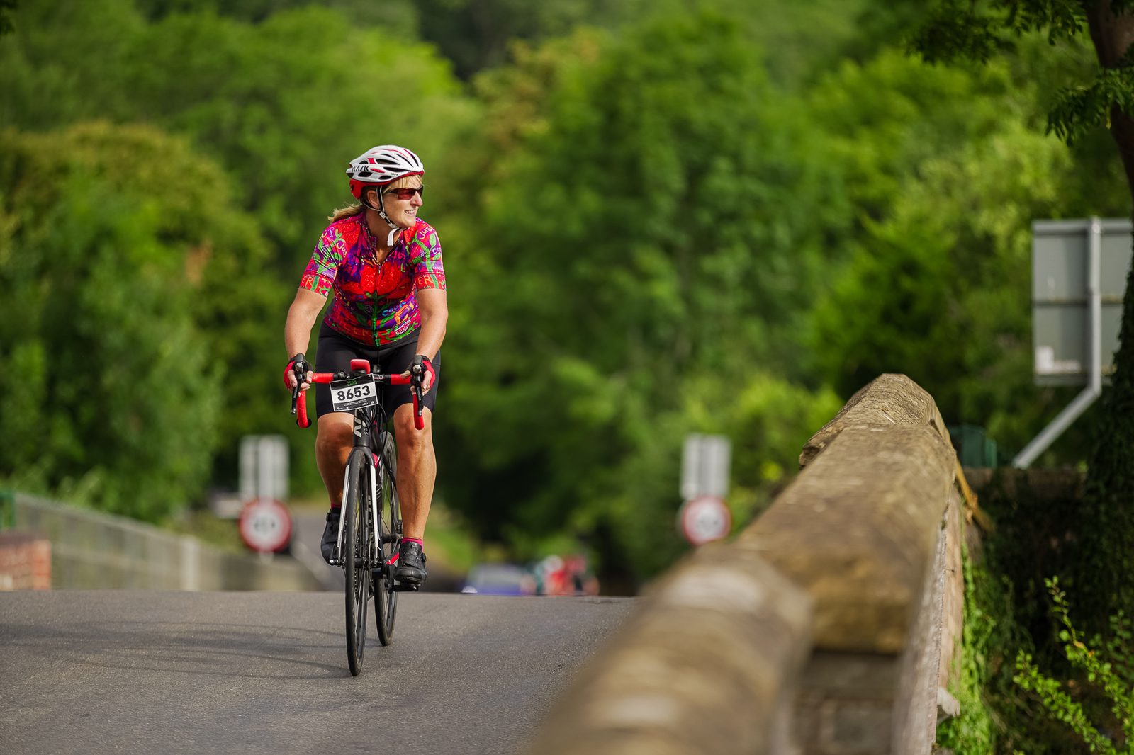 A cyclist wearing a colorful shirt and helmet rides on a road surrounded by lush greenery. The rider appears focused and determined. The background includes a bridge and some distant road signs. The cyclist's bib number is visible as 8635.
