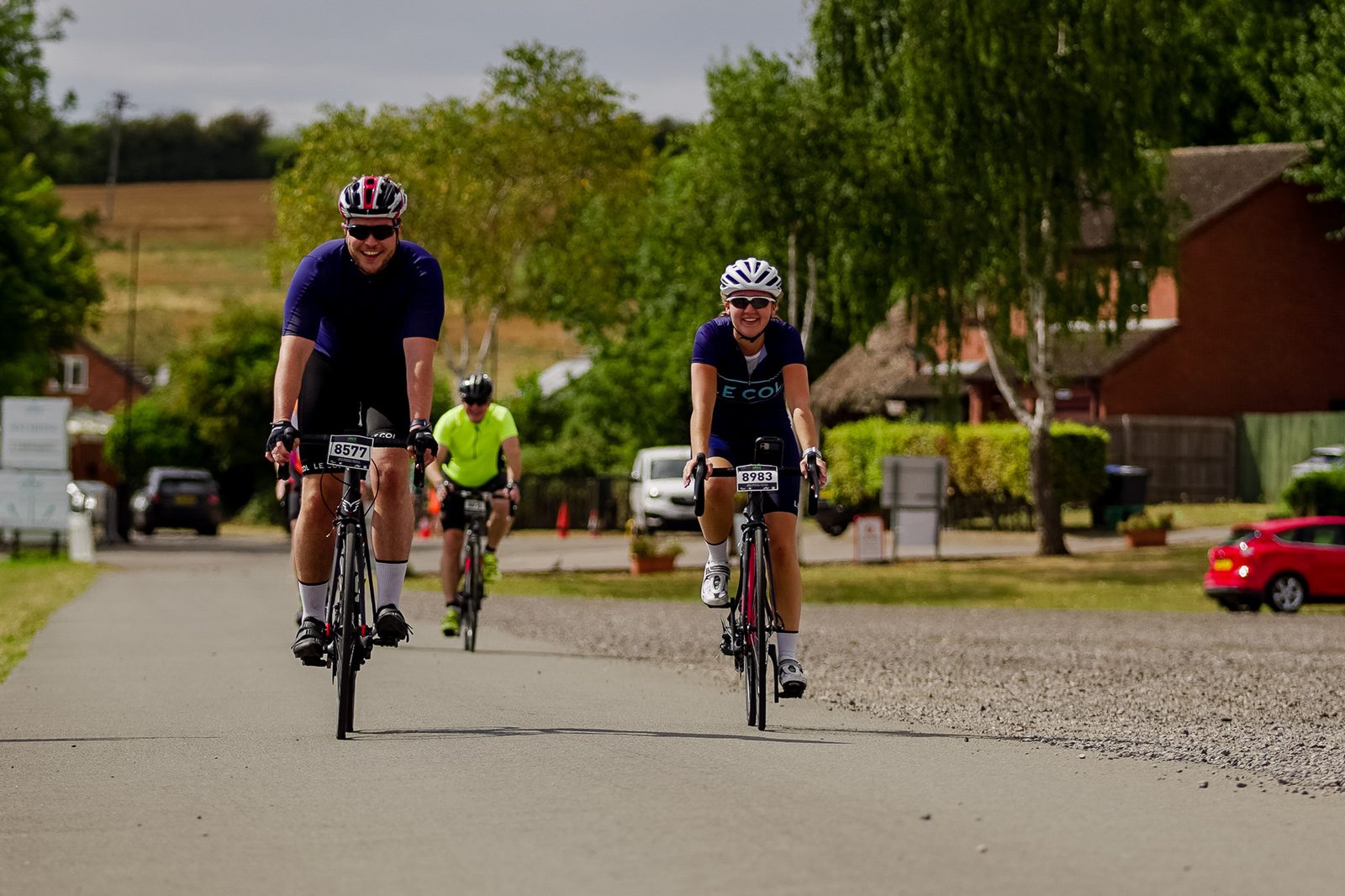 Two cyclists are riding on a paved road in a rural area during the day. A man in a purple shirt and a woman in a navy shirt are in the foreground, both wearing helmets and sunglasses. Another cyclist and a red car are visible in the background.