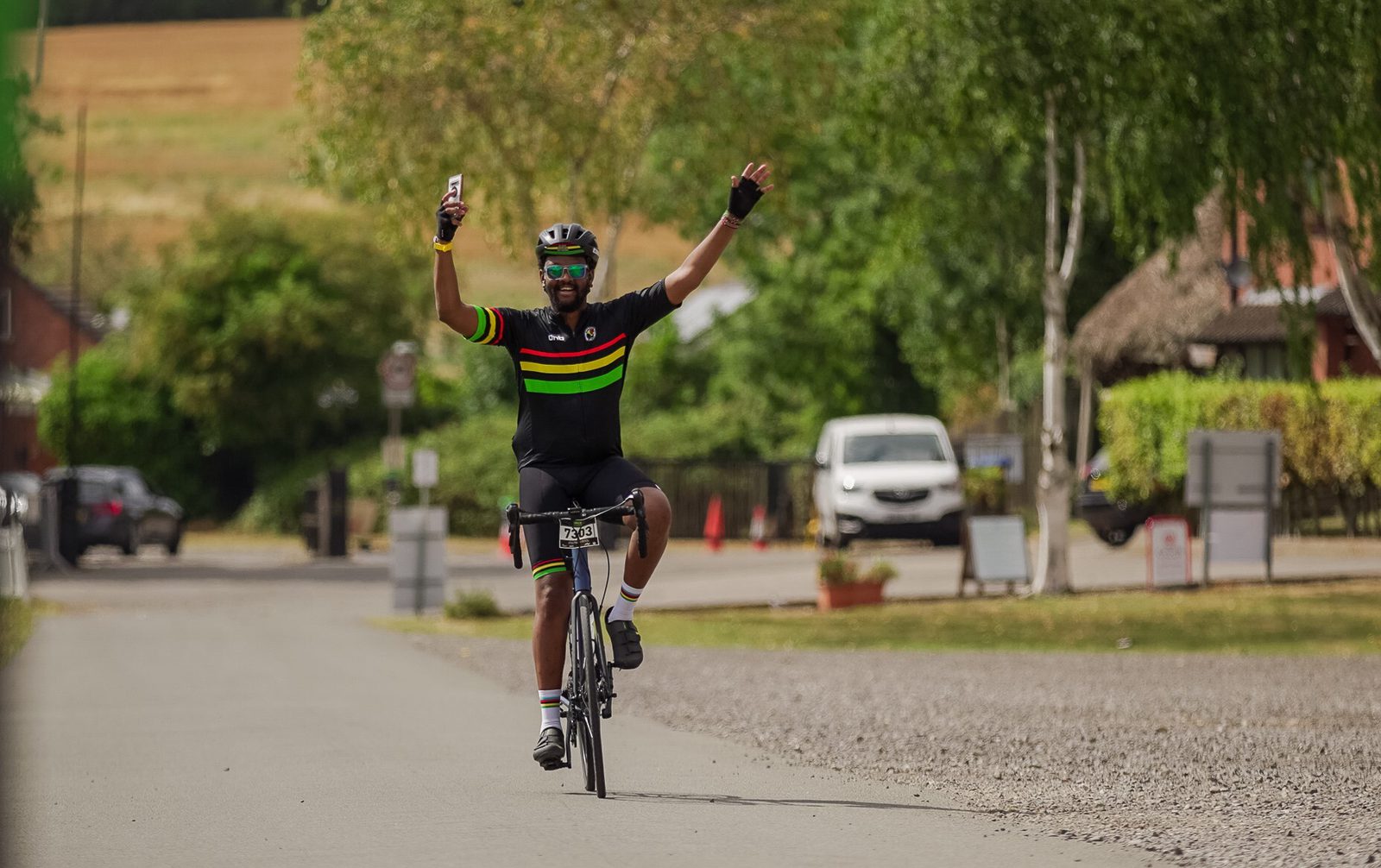 A cyclist wearing a black outfit with colorful stripes raises one arm and holds a phone in the other while riding on a paved path lined with trees and parked cars in the background. The cyclist appears to be smiling and celebrating.