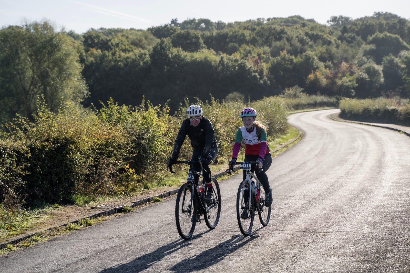 A man and a woman are cycling on a winding road with lush green trees and shrubs on either side. Both are wearing helmets and cycling gear; the woman’s gear is pink and black. The road and the surrounding landscape are brightly lit by the sun.
