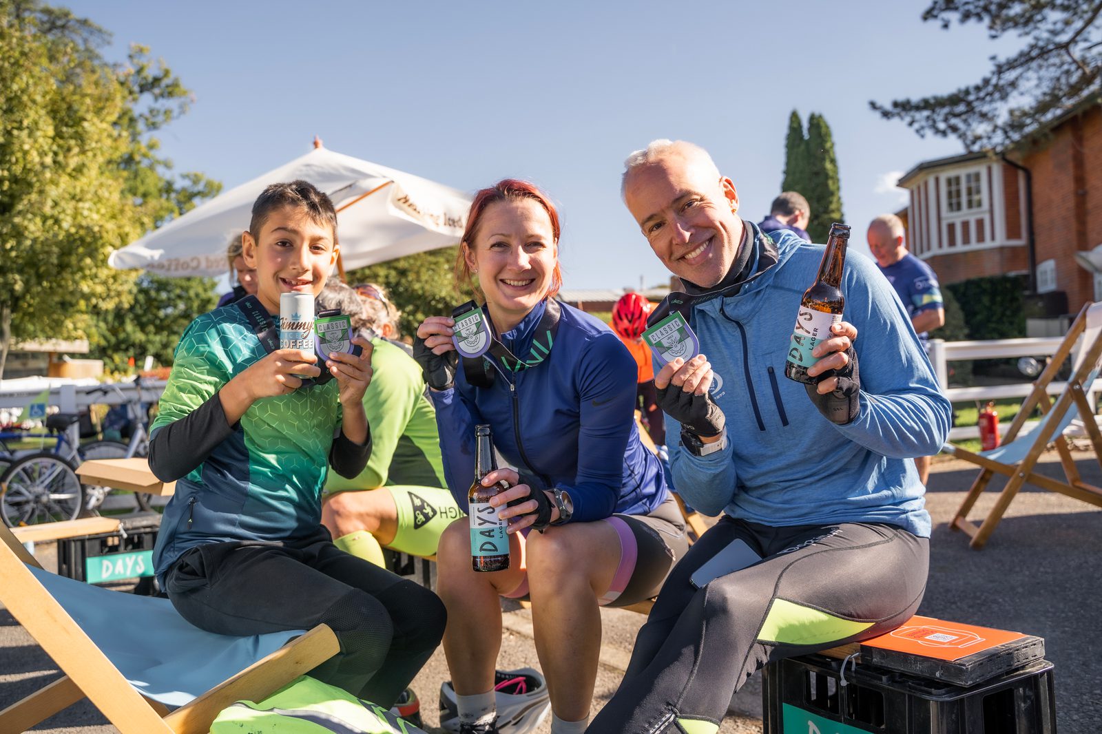Three cyclists, two adults and a child, smile and hold up bottles of non-alcoholic beer while sitting outdoors on a sunny day. They are wearing cycling gear and sitting next to bicycles and outdoor seating, with trees and buildings in the background.