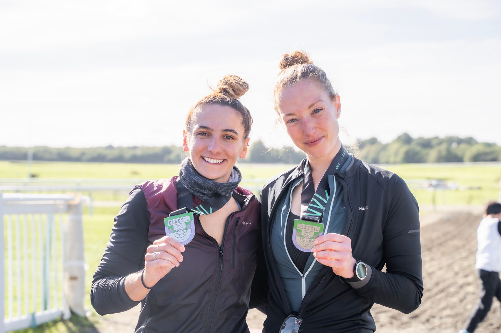Two women stand side by side, smiling and holding medals in an outdoor setting. Both have hair tied in buns and are dressed in athletic wear. A racetrack and field are visible in the background, with bright, natural light illuminating the scene.