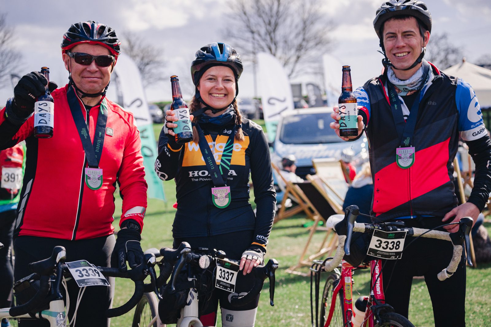 Three cyclists, wearing helmets and medals, stand beside their bikes holding up bottles of drink. They are outdoors with other cyclists and tents visible in the background. The cyclists are smiling and appear to be celebrating their achievement.