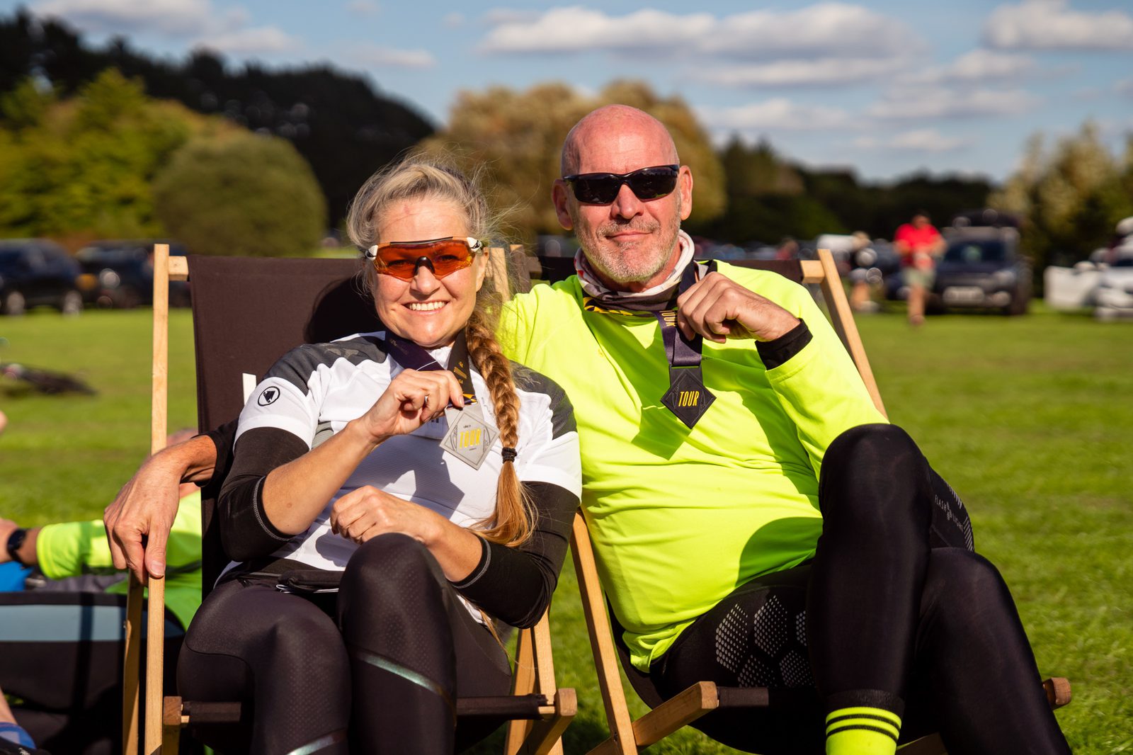 A woman and a man, both wearing sporty attire and sunglasses, sit on deck chairs in a grassy field. They appear to be celebrating, each holding up a medal. The backdrop includes trees, a blue sky with clouds, and parked cars in the distance.