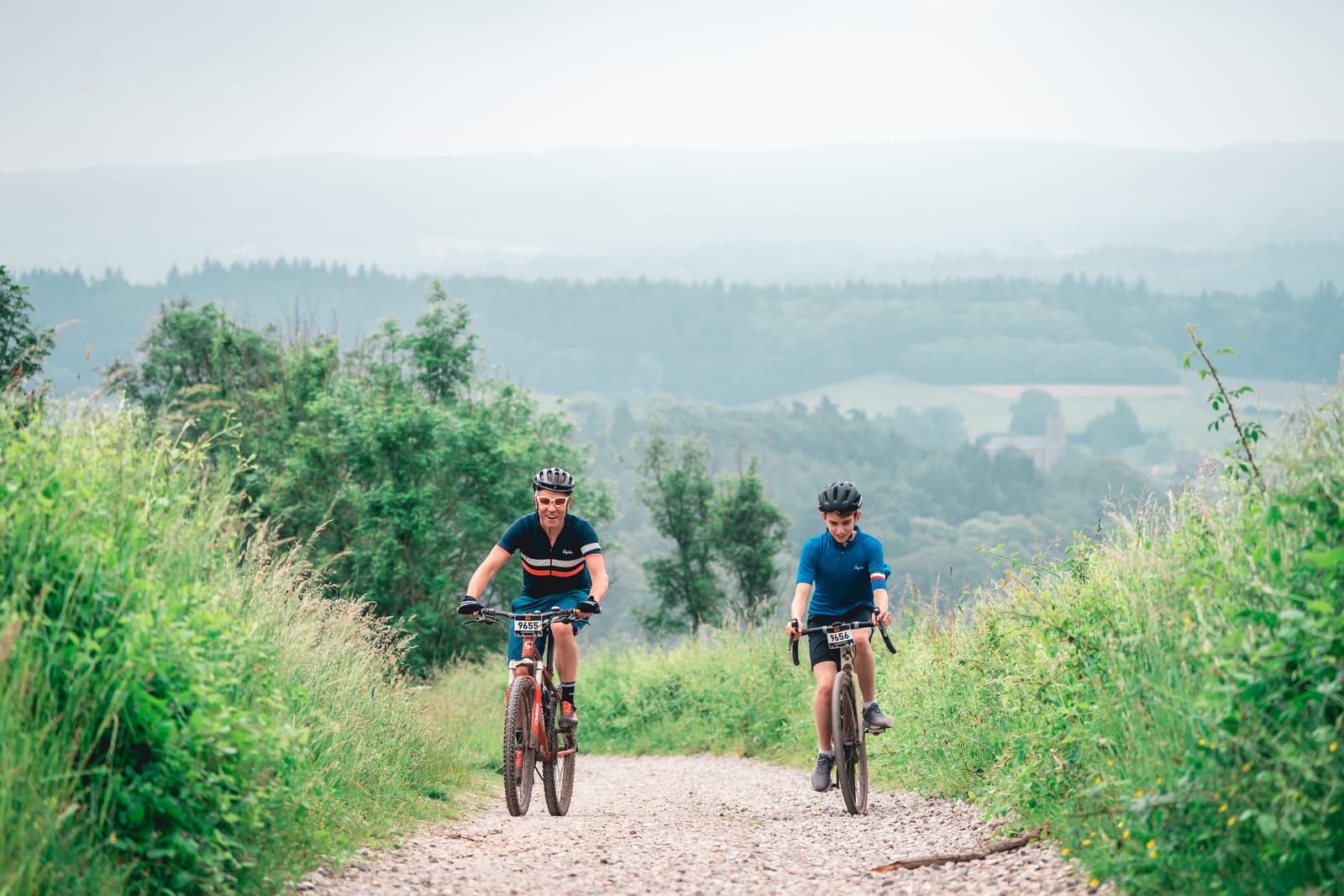 Two cyclists ride mountain bikes along a gravel path surrounded by lush greenery. The background features rolling hills covered in trees, under a cloudy sky. Both cyclists wear helmets and casual attire, enjoying their outdoor adventure.