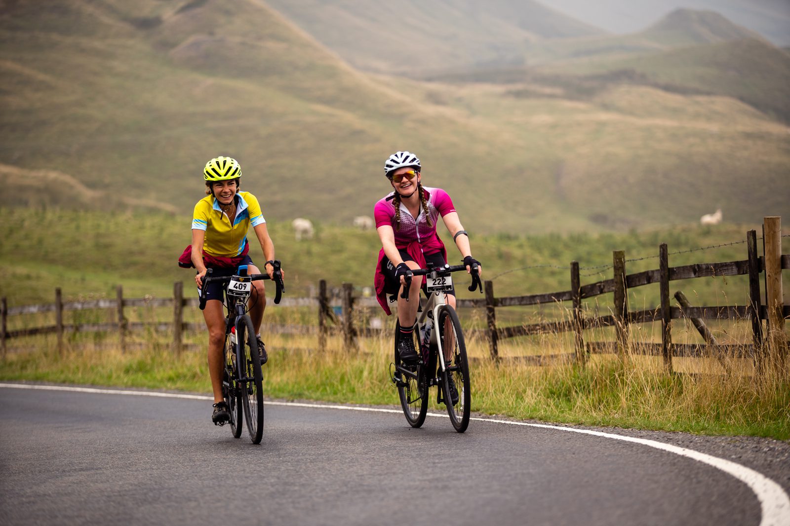 Two cyclists, wearing helmets and colorful jerseys, are smiling as they ride on a scenic, winding road with wooden fences on both sides. The background features rolling green hills and a cloudy sky. Both cyclists have number plates on their bikes.