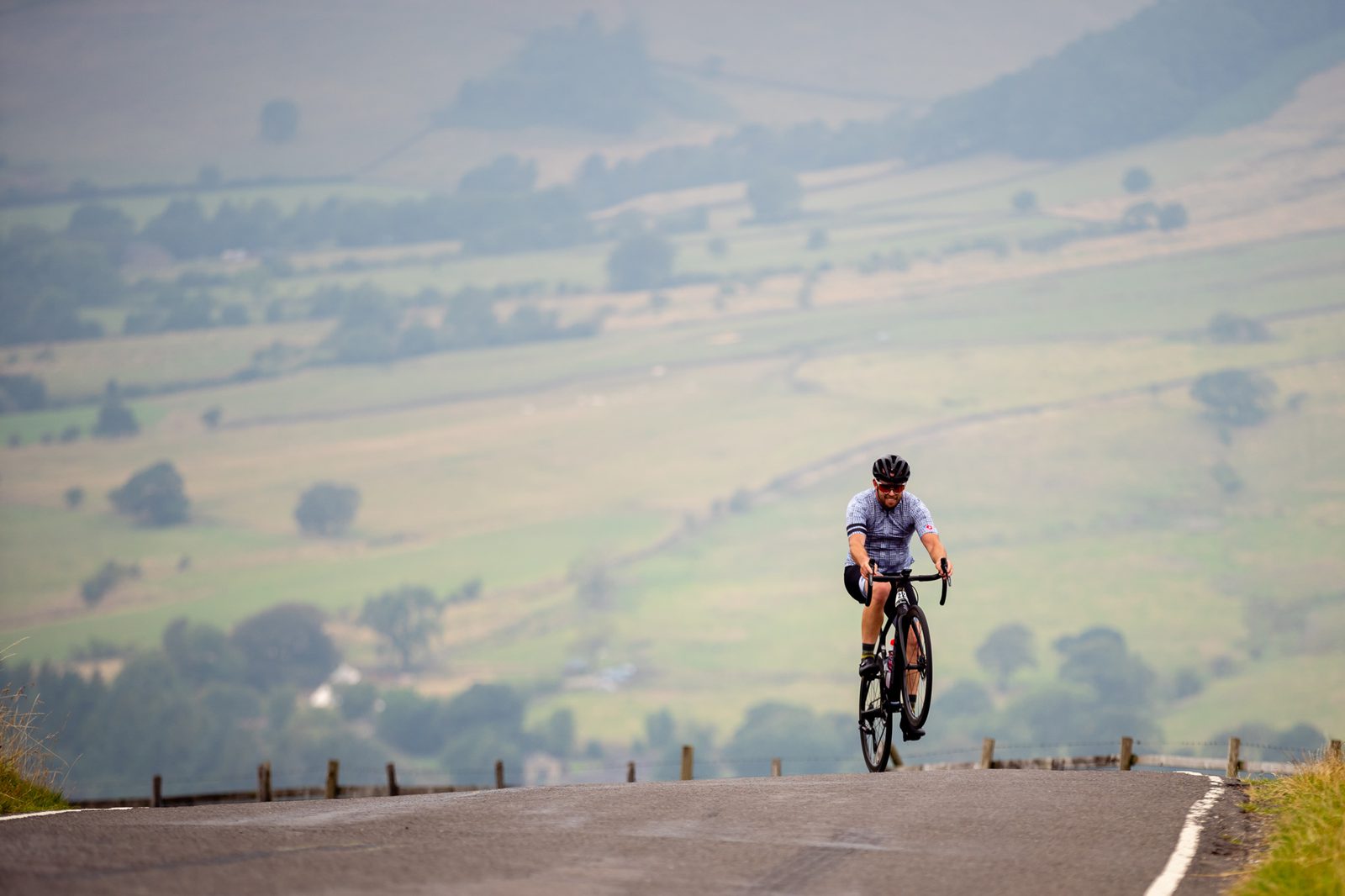 A lone cyclist rides up a paved road on a hilly landscape. The cyclist wears a helmet and a light-colored jersey. The background features an expansive view of rolling green hills and scattered trees under a hazy sky.