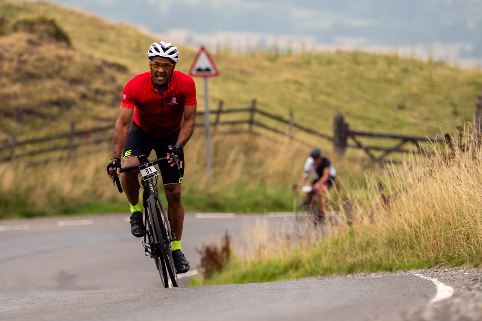 A cyclist in a red shirt, black shorts, and a white helmet is riding up a hilly road, smiling. Another cyclist can be seen in the background. The road is flanked by grassy hills and a wooden fence, with a traffic sign visible in the distance.