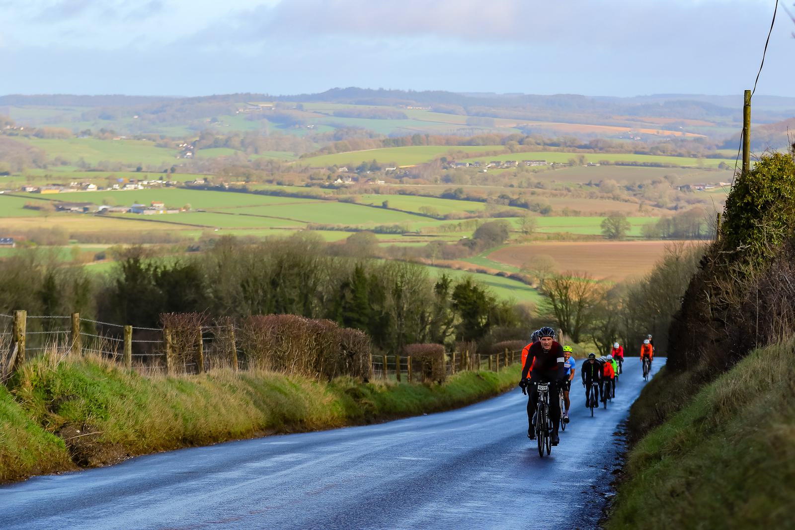 A group of cyclists rides down a rural road surrounded by lush greenery and rolling hills on a sunny day. The landscape features fields, scattered houses, and trees under a partly cloudy sky. The road is bordered by hedges and has a slight incline.