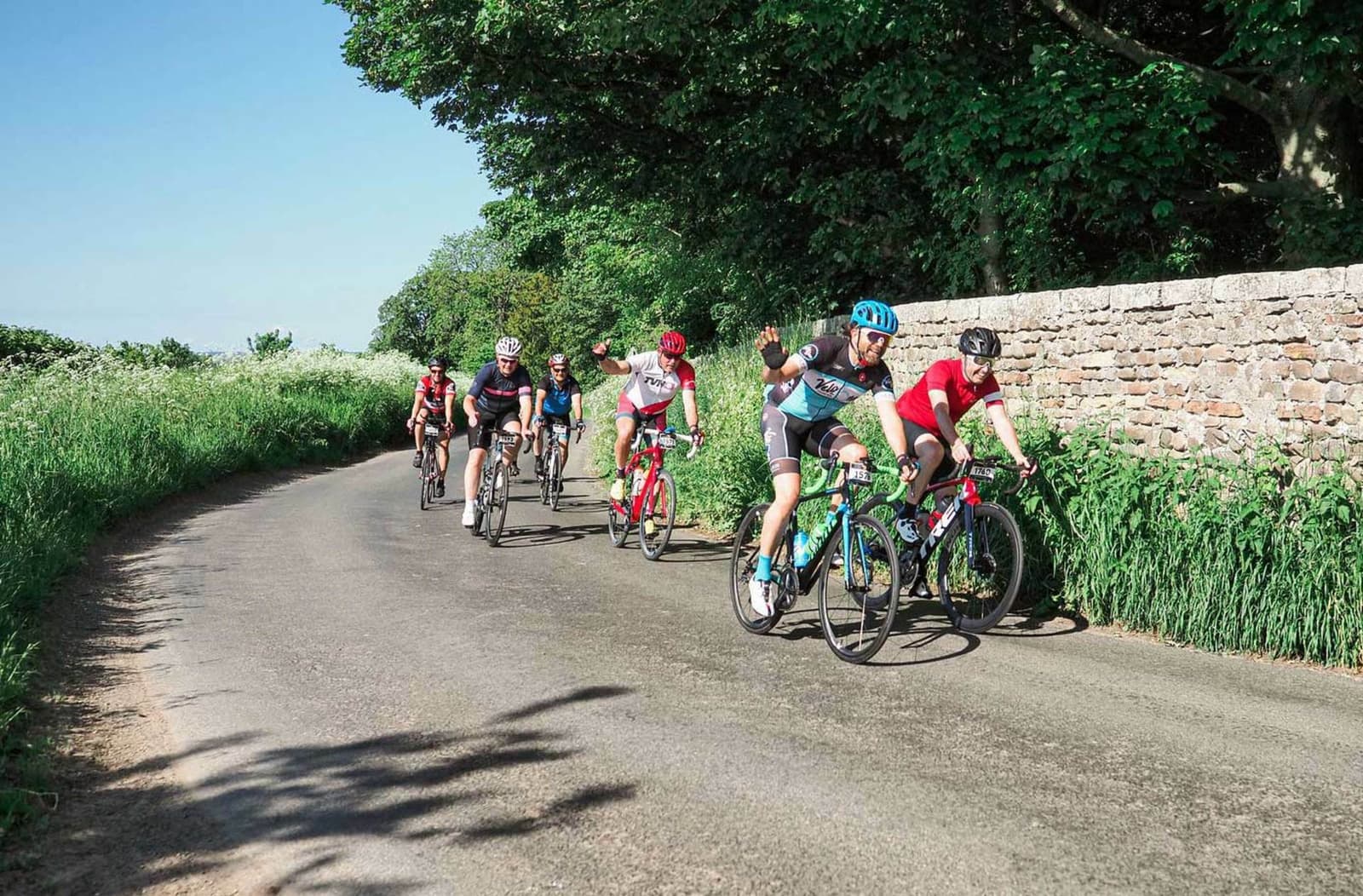 A group of cyclists riding along a rural road bordered by greenery and a stone wall on a sunny day. One cyclist in the foreground is waving at the camera. The scene is vibrant and lively, with trees providing partial shade.