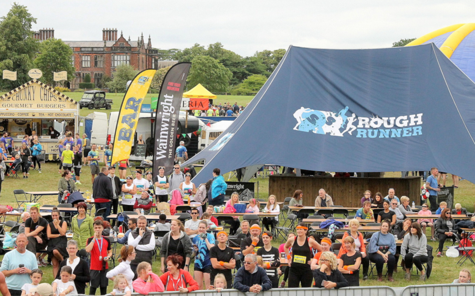 A large outdoor event with many people gathered around tables and tents. A blue "Rough Runner" tent is prominent in the center, with other flags and food stalls in the background. A historic building and trees are visible in the distance.