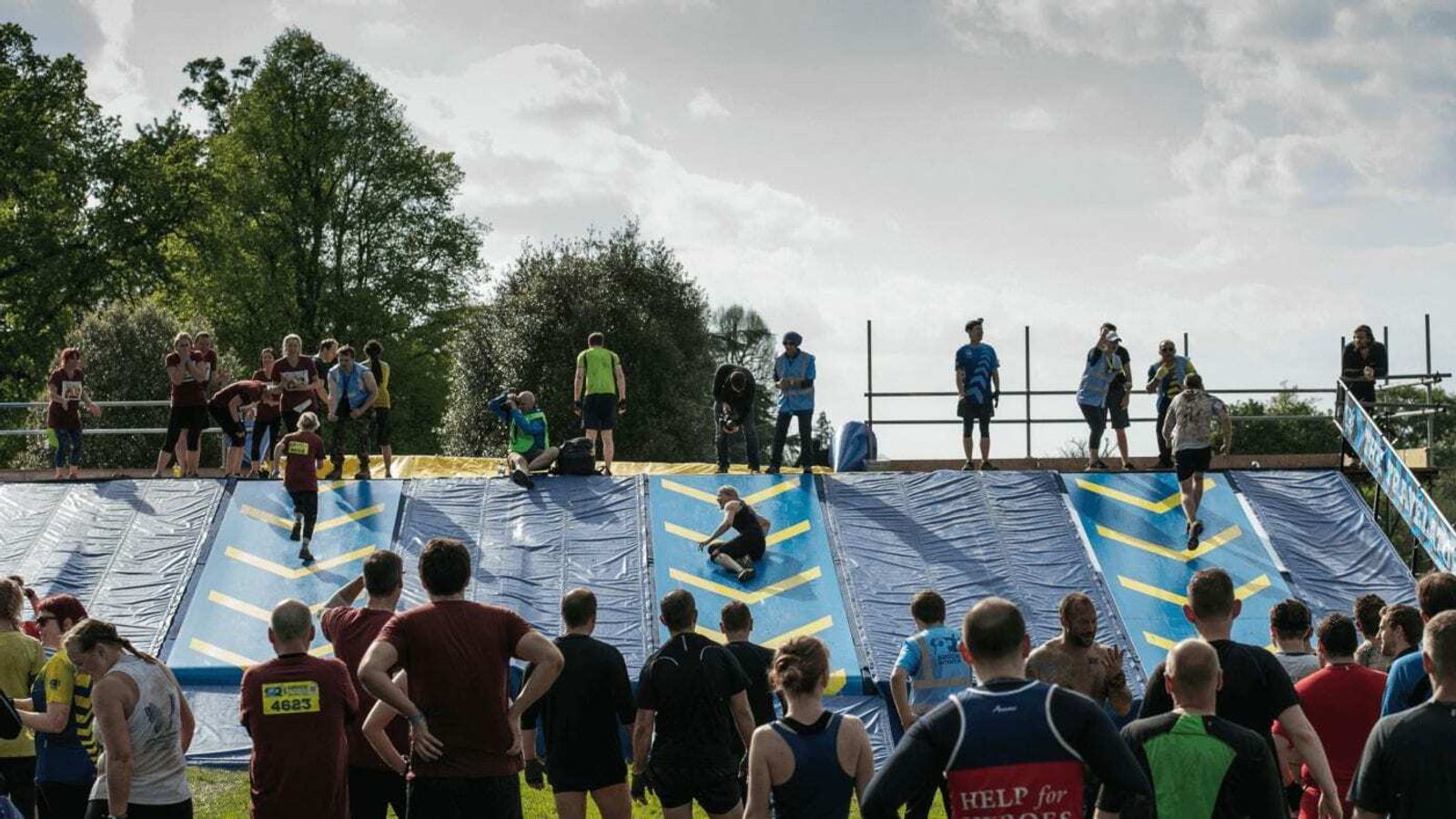 Participants are climbing a steep, slippery wall adorned with yellow grips during an outdoor obstacle course race, while spectators watch from below. The setting appears to be a park, with trees and a cloudy sky in the background.