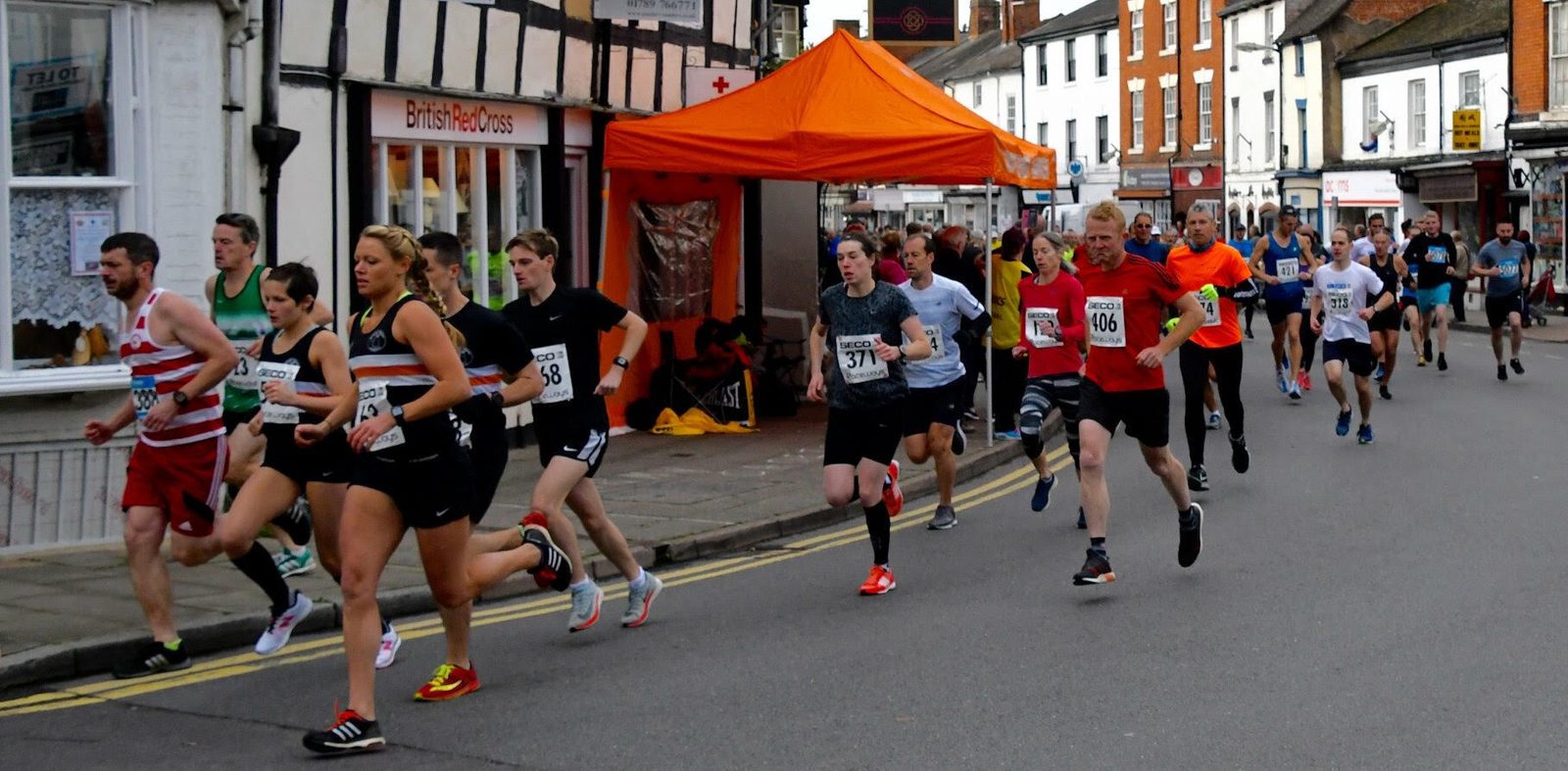 Runners participate in a street race, passing buildings and an orange tent. Some wear race bibs with numbers like 108, 377, and 140. The atmosphere appears lively, with participants of various ages and fitness levels. The background includes quaint storefronts.