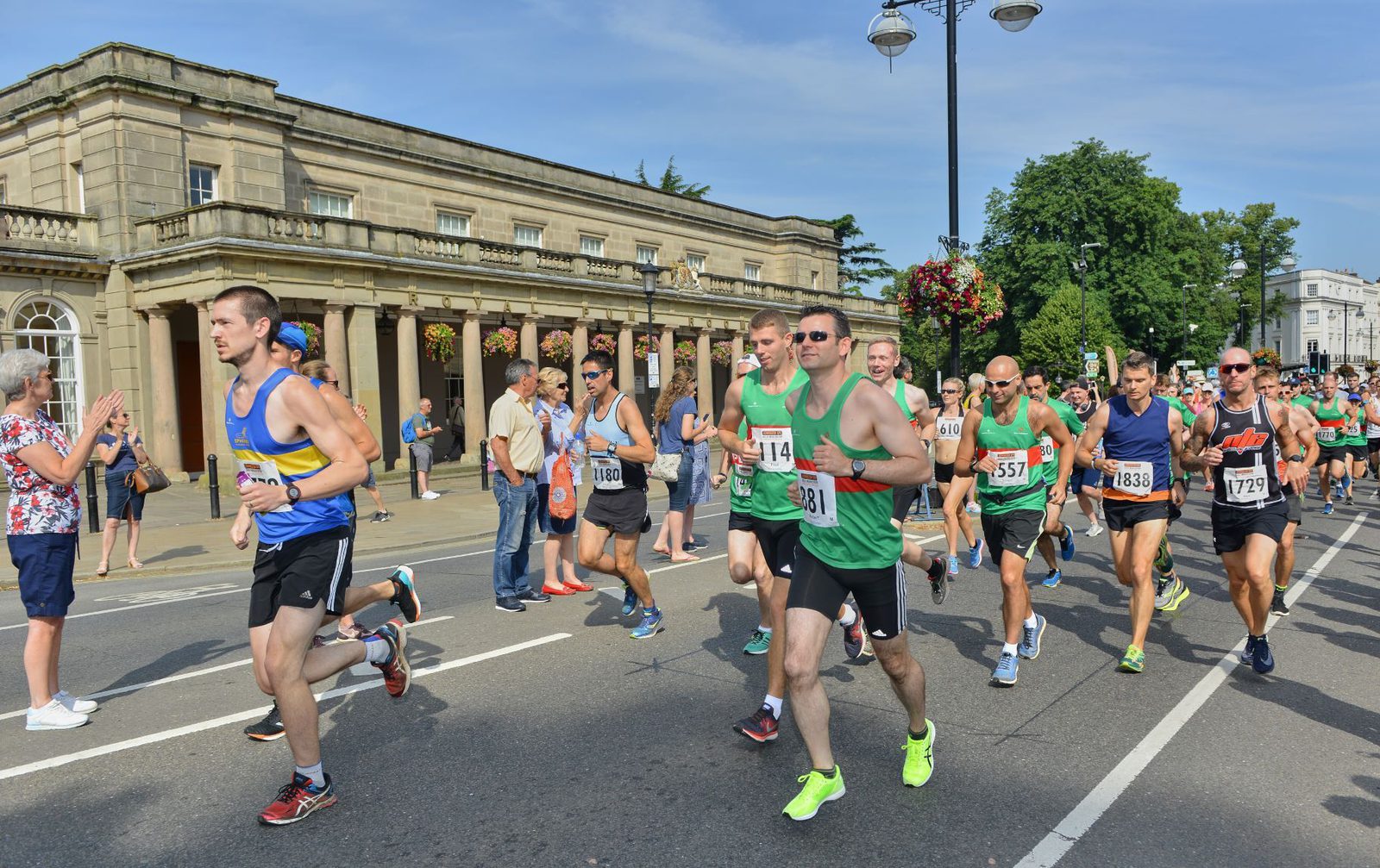 A group of runners in colorful athletic wear compete in a marathon on a sunny day. They run past a historic building with columns and arched windows. Spectators stand on the sidelines, cheering and clapping. The atmosphere is lively and energetic.