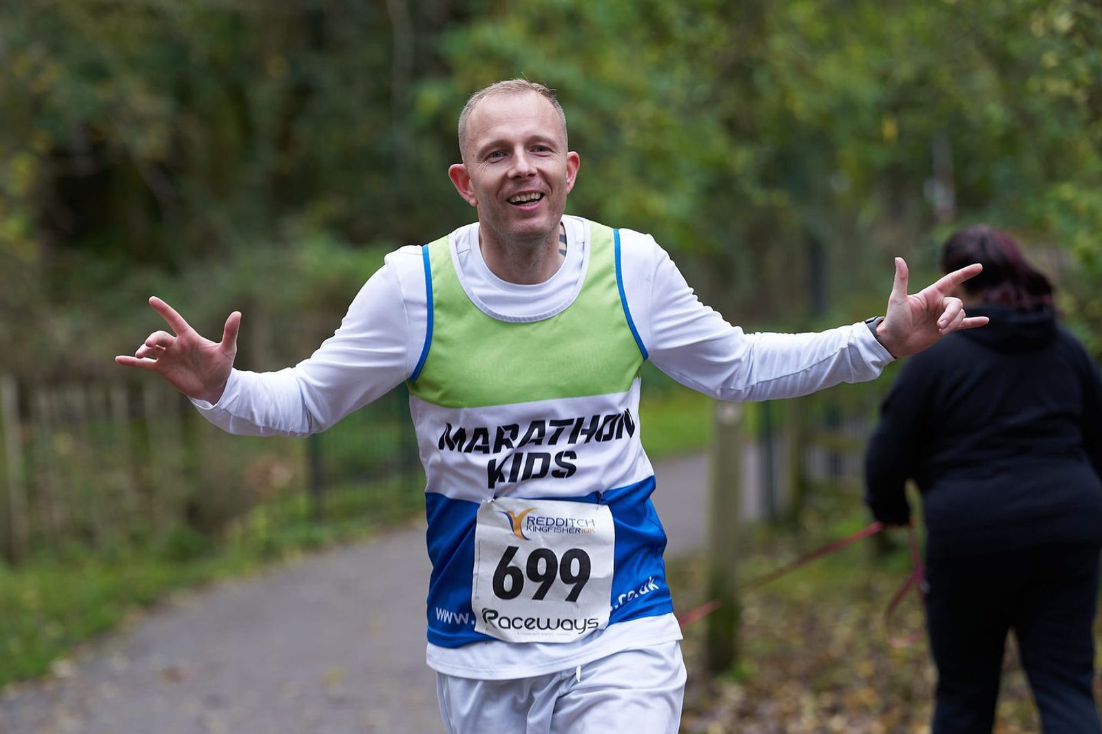 A smiling runner wearing a green and white "Marathon Kids" shirt and bib number 699 gestures with both hands while jogging on a trail surrounded by greenery. Another person can be seen slightly blurred in the background with their back turned.