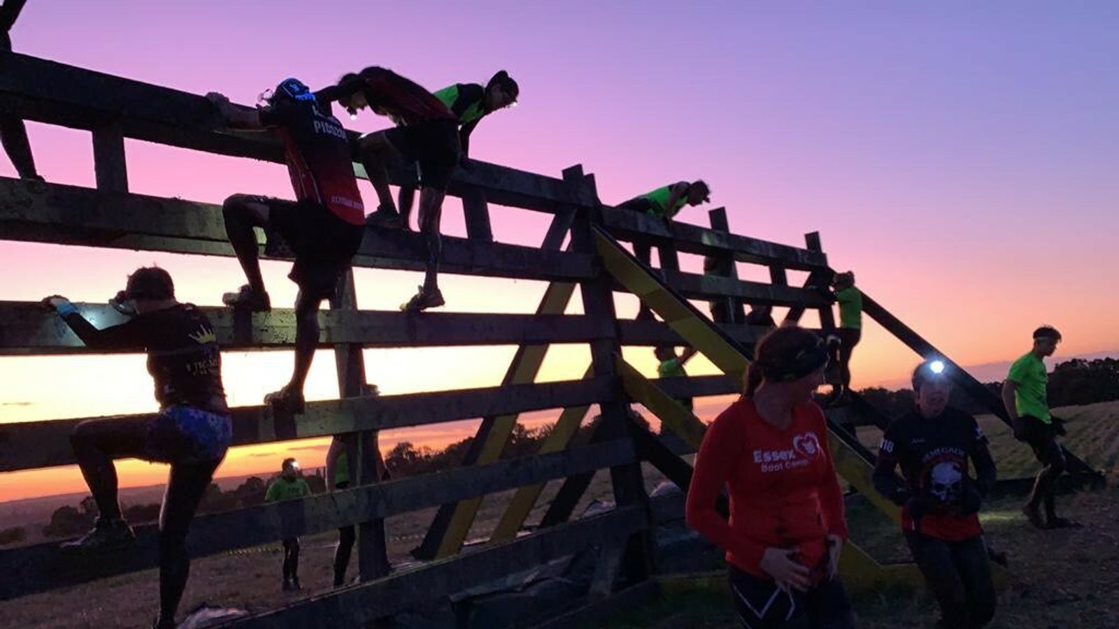 People in athletic gear, some illuminated by headlamps, are climbing over a large wooden obstacle at dusk. The sky is a gradient of purple and orange, indicating the setting or rising sun. The terrain appears rugged and natural, enhancing the challenge.