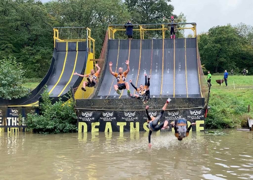 A group of people in colorful athletic clothing are sliding down a series of steep, wet ramps and launching off into a muddy water pit below, arms and legs flailing in mid-air. The backdrop features green trees and a cloudy sky. The scene is lively and energetic.