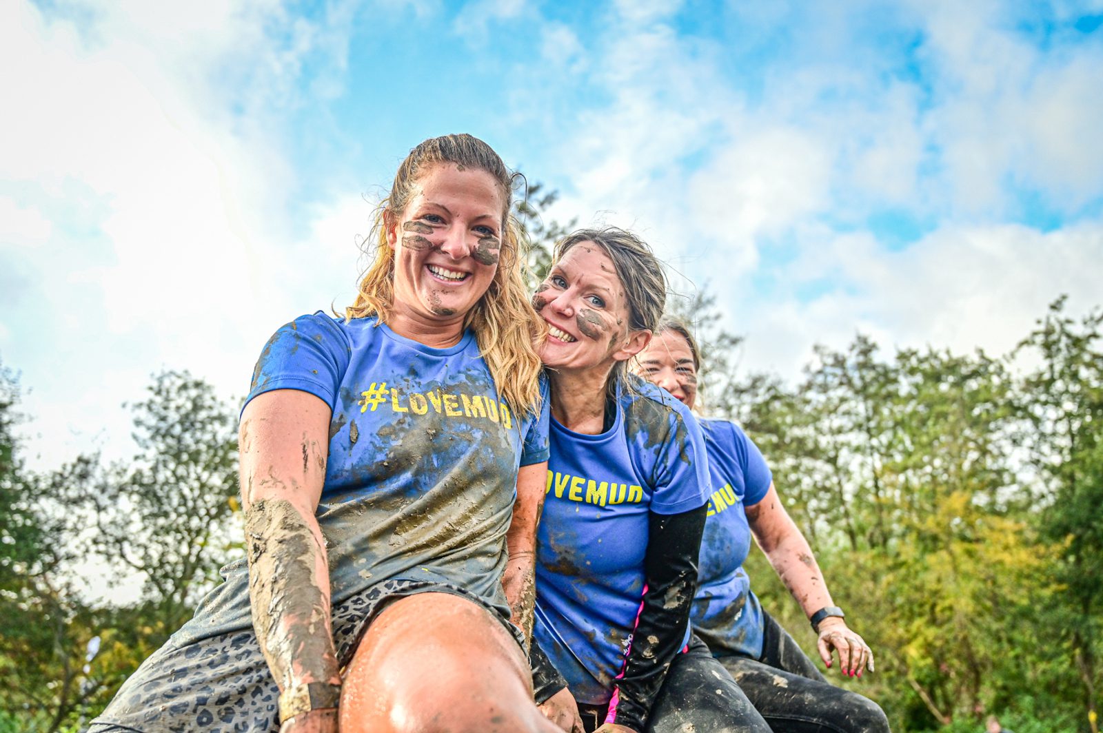 Three women covered in mud, wearing blue shirts with "#LOVEMUD" printed on them, are smiling and posing together outdoors. The background shows trees and a blue sky with scattered clouds, indicating they are participating in an outdoor mud run or obstacle course.