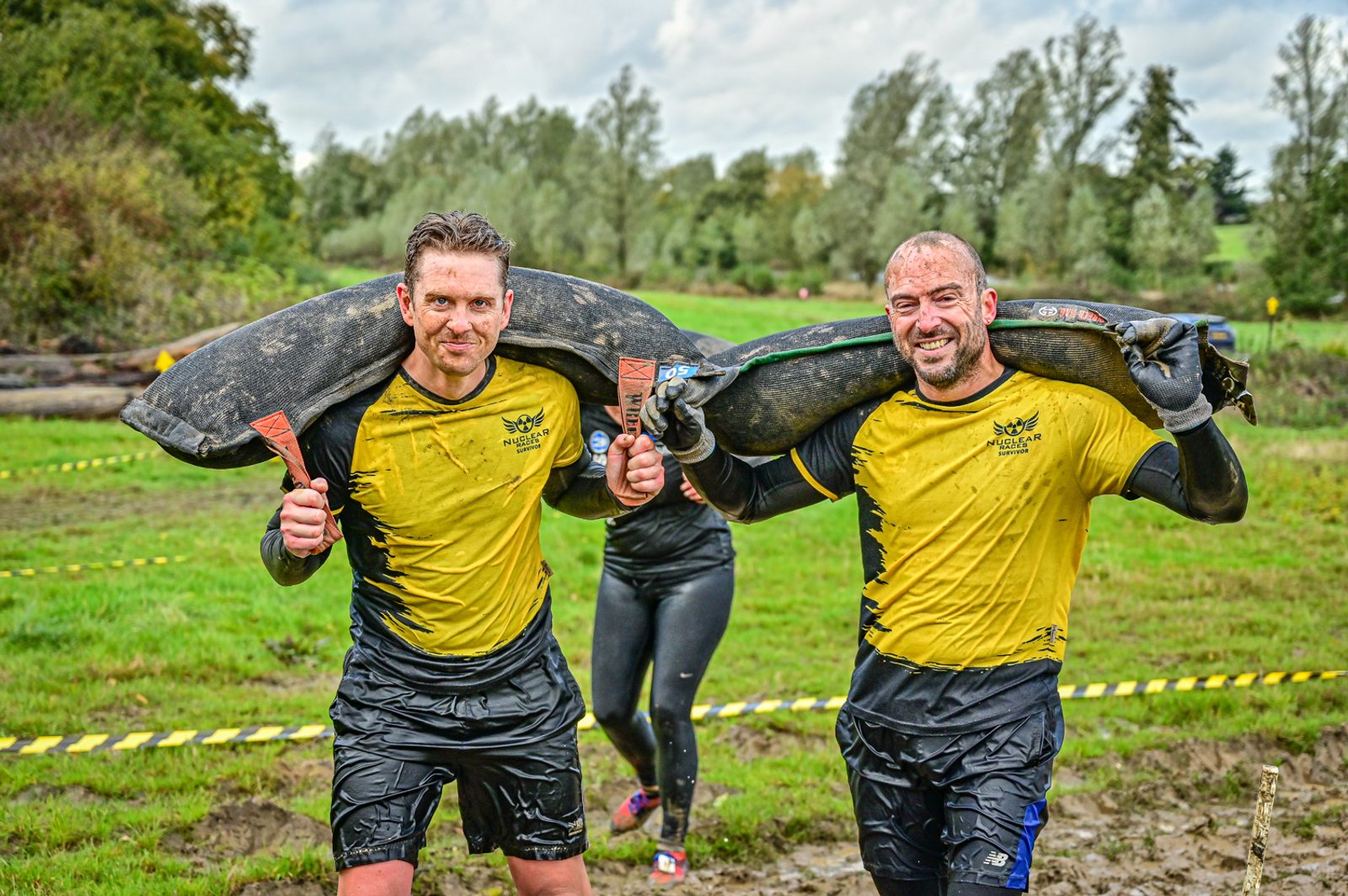 Two men wearing yellow shirts carry heavy, muddy sandbags over their shoulders during an outdoor obstacle course event. The men, covered in mud, are smiling and walking through a grassy field with trees and cloudy skies in the background.