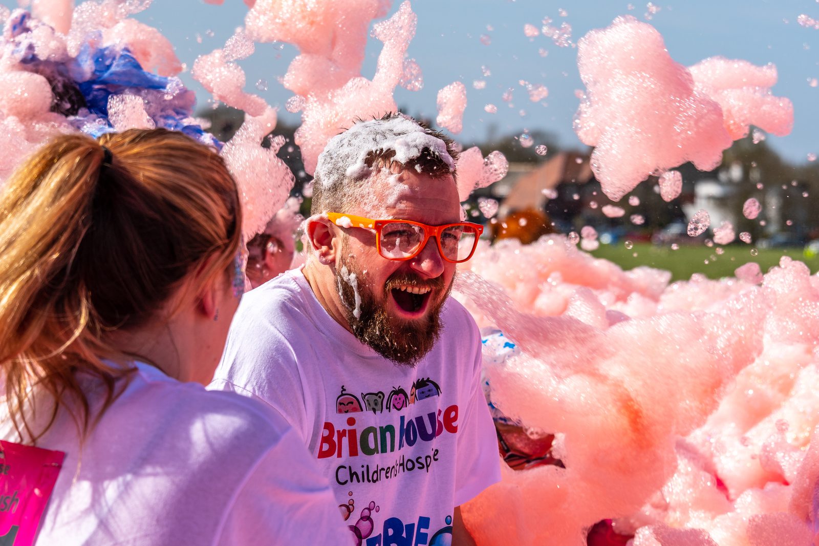 A man with a beard and red-framed glasses laughs as he is covered in pink foam during an outdoor event. He is wearing a white t-shirt with "Brianhouse Children's Hospice" printed on it. Another person nearby is also covered in foam. The sky is clear and blue.