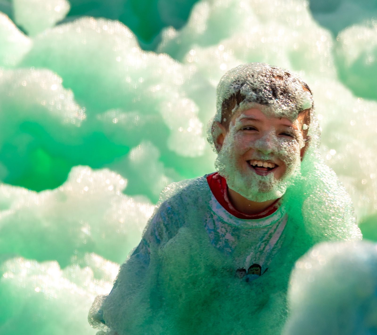 A young child with a foam-covered face is smiling joyfully, standing amidst thick green and white foam. The child is wearing a red shirt under a white top, with light reflecting off the bubbles, creating a fun and playful atmosphere.