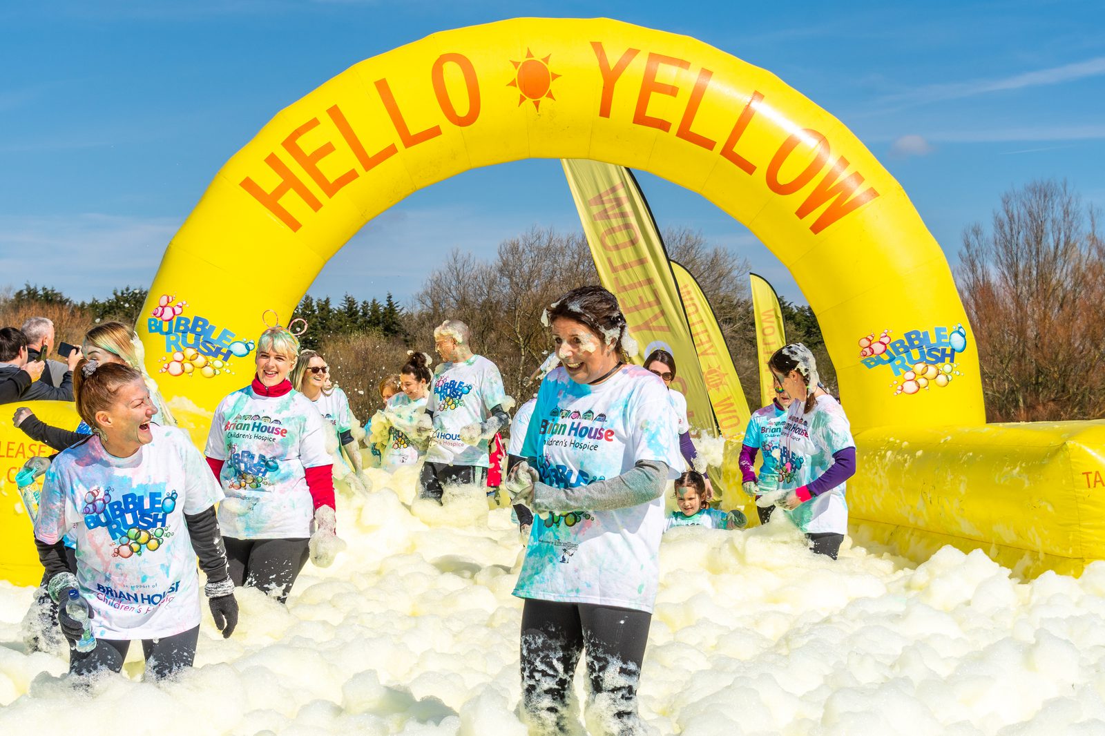 Participants, covered in foam, smile and laugh as they walk through a thick layer of bubbles under a bright yellow arch reading "HELLO YELLOW" at a bubble run event. They are wearing white event T-shirts and the sunny sky is clear in the background.