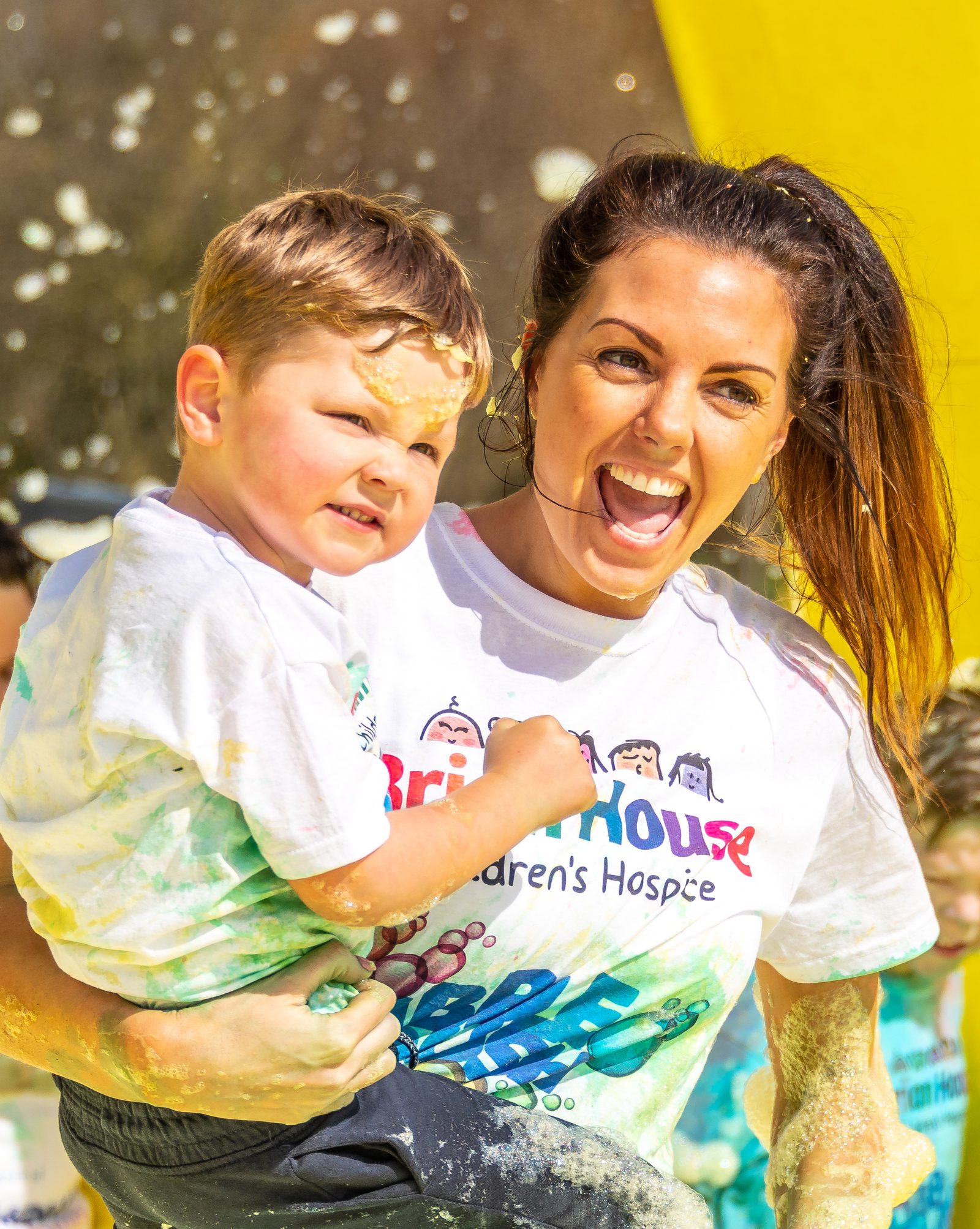 A woman with brown hair in a ponytail joyfully holds a smiling young boy while both are covered in paint splashes. They are wearing white t-shirts with colorful designs and text that includes "Children's Hospice." The background is a vibrant, sunny outdoor setting.