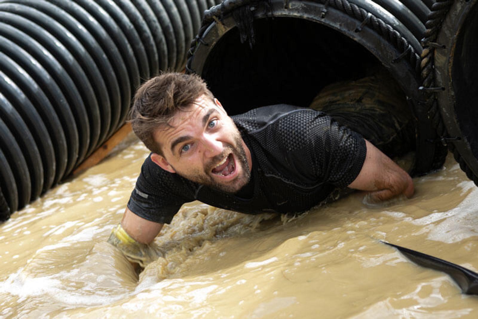 A man emerges from a muddy water-filled tunnel, wearing a black shirt and gloves. He appears excited or determined, with water splashing around him as he moves forward in the obstacle course.