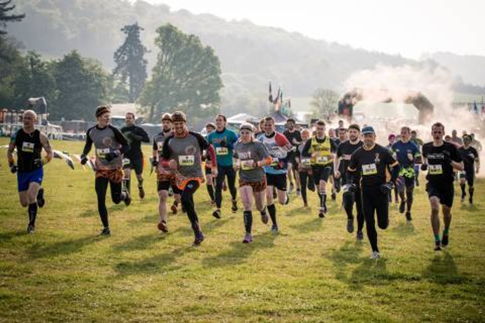 A group of runners, dressed in various athletic outfits, race across a grassy field. The background features trees, hills, and a misty or smoky area near the starting point. Some participants appear determined while others seem to be enjoying the event.