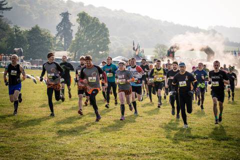 A group of runners, dressed in various athletic outfits, race across a grassy field. The background features trees, hills, and a misty or smoky area near the starting point. Some participants appear determined while others seem to be enjoying the event.