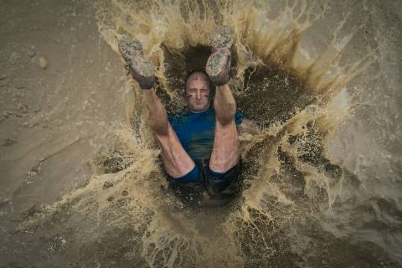 A person in muddy clothes is captured mid-air as they fall into a large mud puddle, creating a dramatic splash with legs raised and arms just submerged in the water. The surrounding area appears wet and muddy.