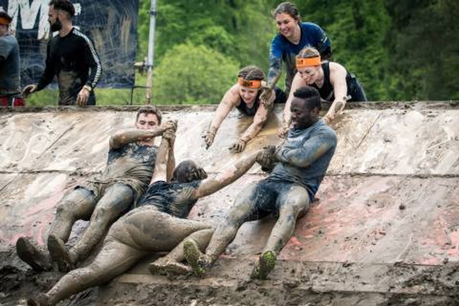 Participants in a tough mudder event are covered in mud, working together to climb a slick, inclined wall. Some are pulling others up, showcasing teamwork and determination. The background features greenery and observers cheering them on.