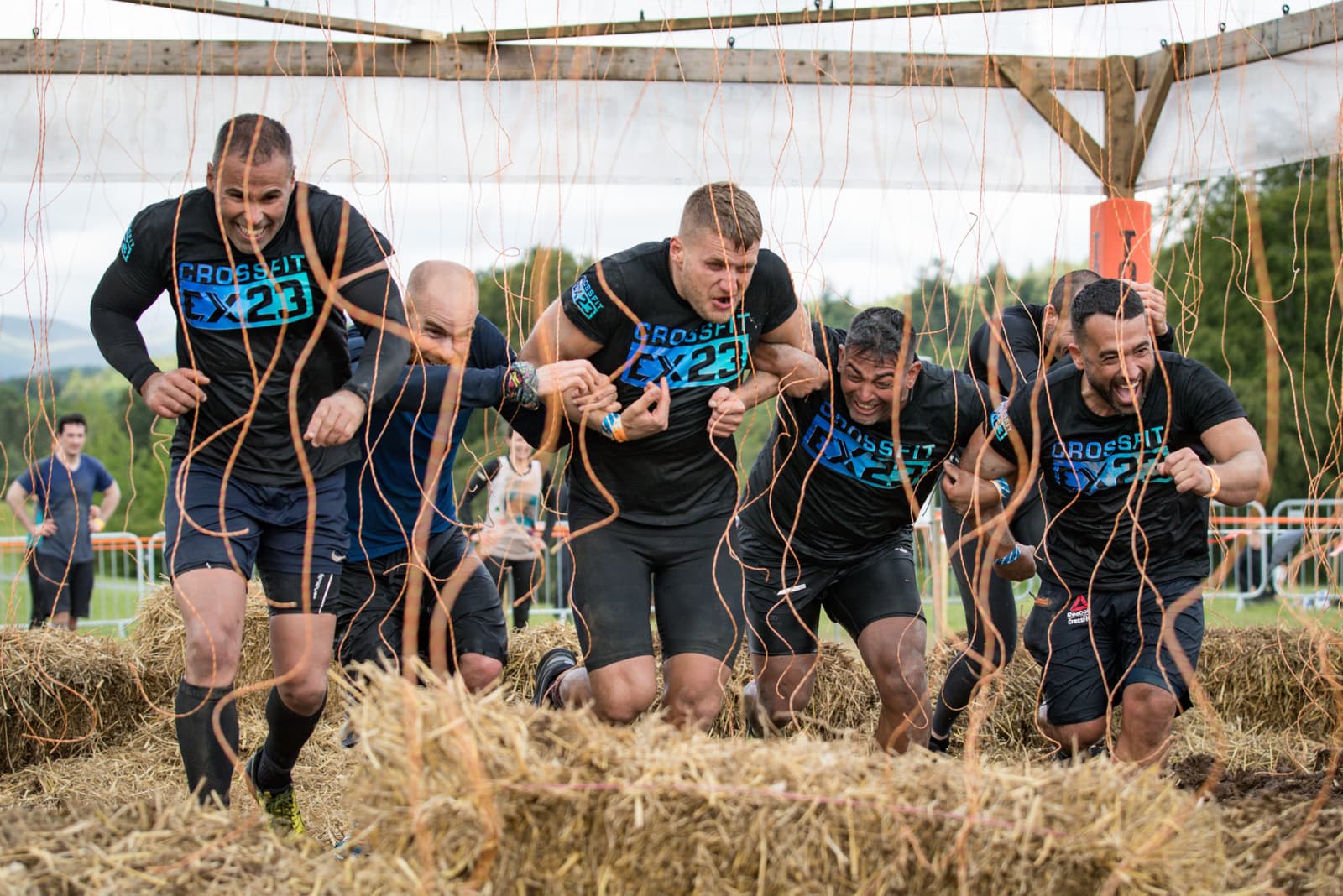 A group of six people in black CrossFit shirts navigate through an obstacle course with hanging wires, surrounded by hay bales. They appear to be running together with focused and determined expressions. The setting looks like an outdoor event in a grassy area.