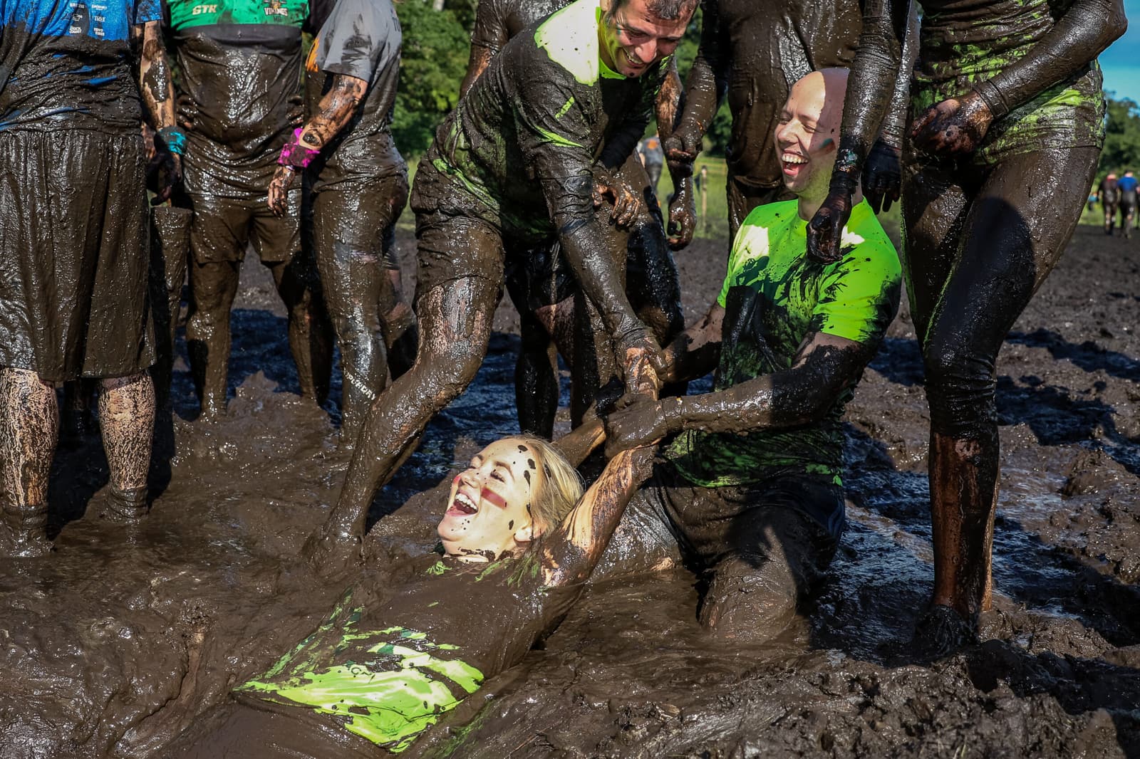 A group of people in athletic clothing, covered in mud, are participating in a muddy obstacle course. One person on the ground is being playfully pulled up by two others, all of them smiling and laughing. The background shows trees and a clear sky.