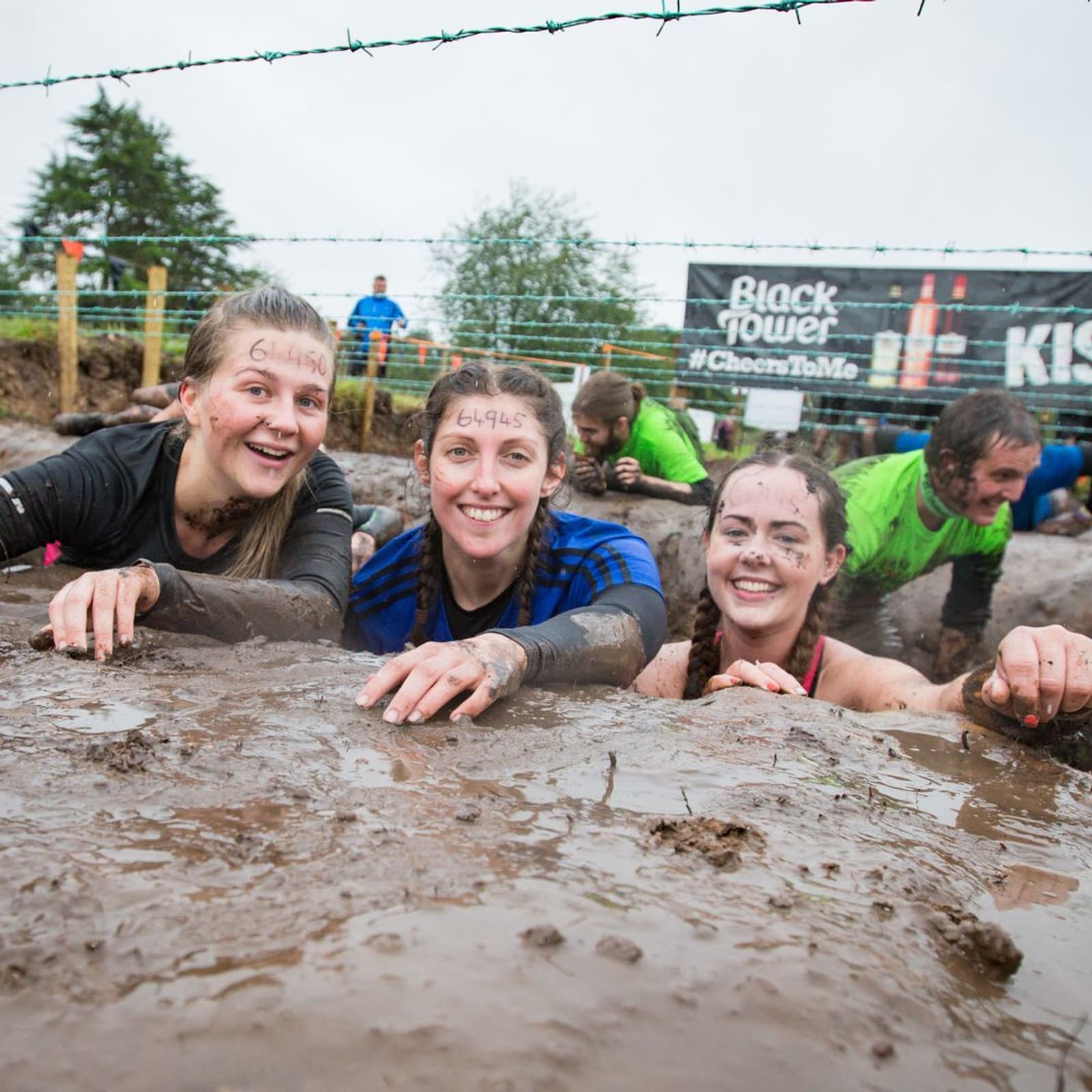 Three smiling women covered in mud participate in an outdoor obstacle course, crawling under barbed wire. They are having fun despite the challenging conditions. Other participants can be seen in the background, along with a banner displaying the text "Black Tower.