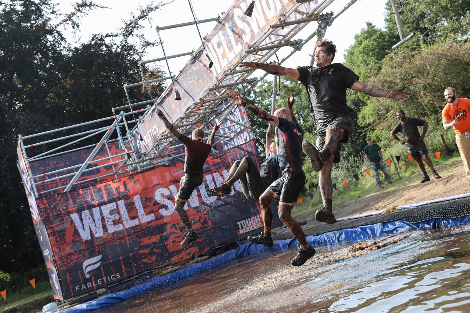Participants in a mud run are attempting to cross a set of monkey bars above a muddy water pit. One person is mid-air after missing a bar, while others are still gripping the bars. A banner and onlookers are visible in the background.