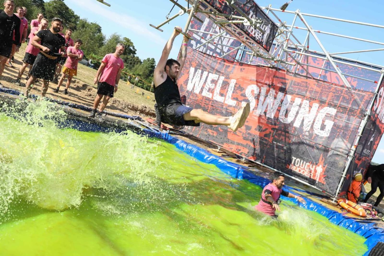 A participant swings from a metal bar over a pool of green water at a Tough Mudder event. Others watch from the edge, and a person in the water encourages the swinger. A banner reading "Well Swung" is visible in the background.